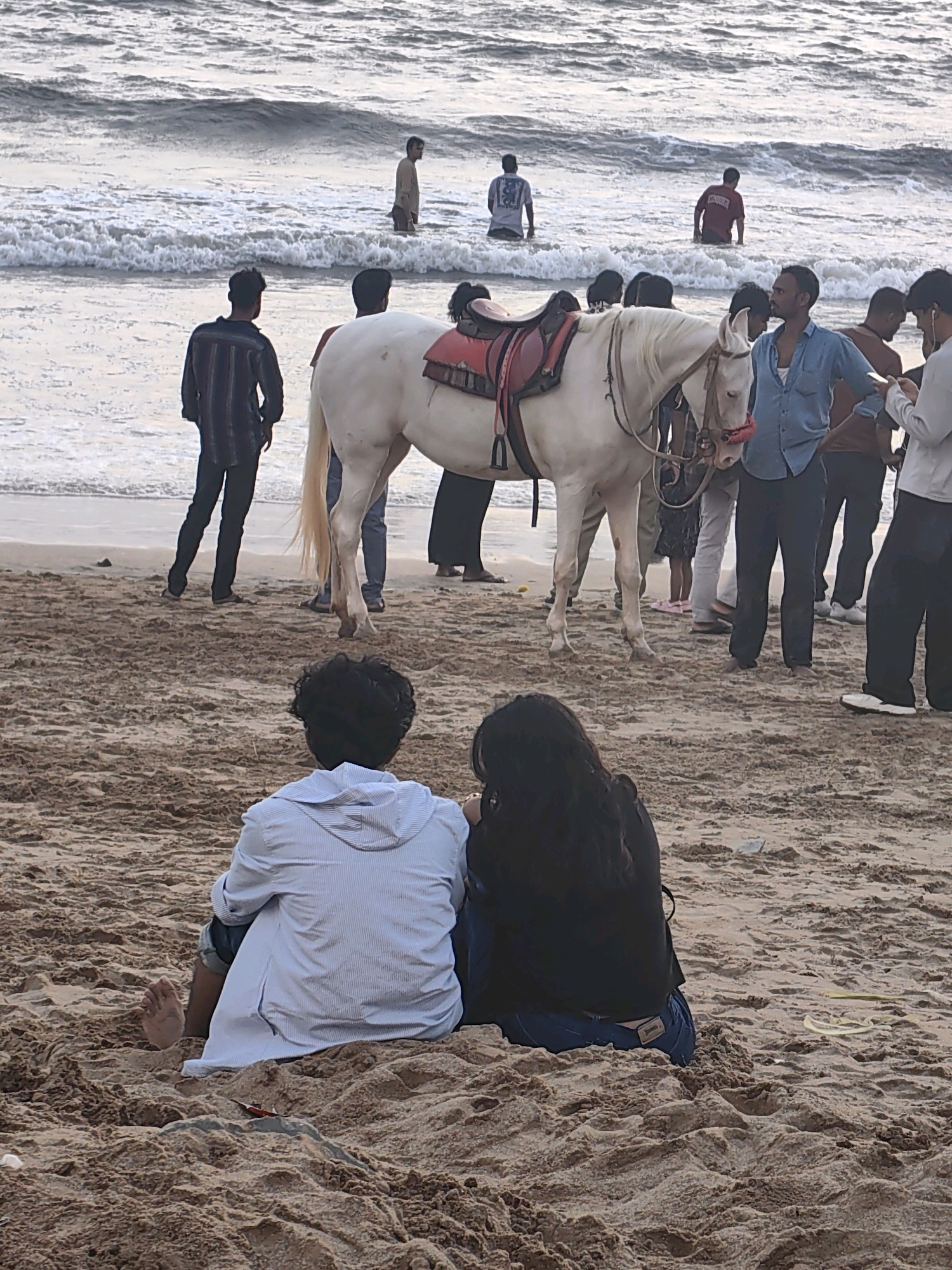 This image depicts a scene at Juhu Beach in Mumbai, India. The foreground shows two individuals, a male and a female, seated on the sand with their backs to the camera, facing the ocean. The male is wearing a striped hooded shirt and jeans, and his bare foot is visible. The female is wearing a black top and jeans.
In the mid-ground, a white horse with a red saddle stands on the wet sand near the water's edge. Several people are gathered around the horse, some appearing to be tourists or locals interacting with it. A man in a blue shirt stands close to the horse's head. Further out in the shallow water, several other individuals are standing or wading, with waves breaking around them. The overall environment is a beach with sandy shores and the Arabian Sea. The lighting suggests it is likely late afternoon or early evening.