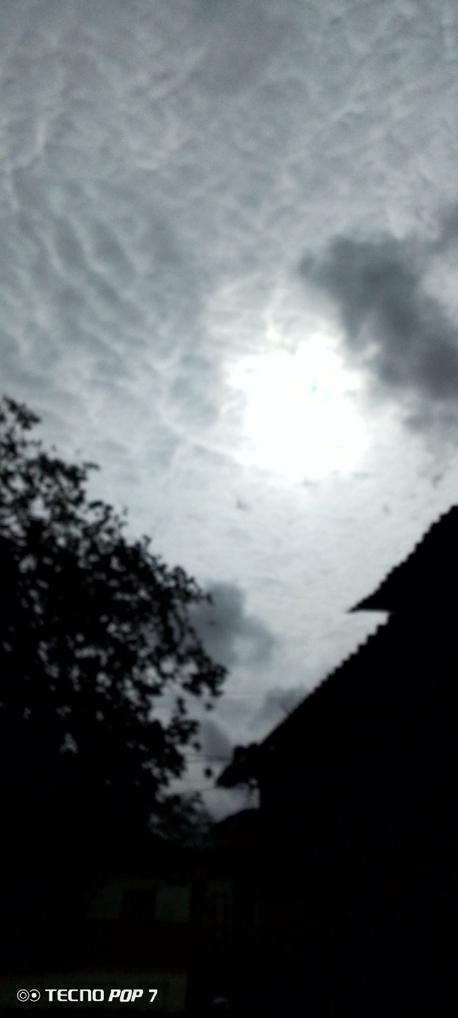 The image is an over-the-shoulder shot taken outdoors, looking upwards at the sky. The sun is partially obscured by clouds, creating a bright, washed-out area in the center of the frame. To the left, the dark silhouette of tree branches and foliage is visible. To the right, the dark silhouette of a roofline with a distinct angled edge cuts across the frame from the bottom right corner towards the upper center. The sky is filled with a pattern of streaky, textured clouds, with darker clouds clustered around the sun. The overall lighting suggests an overcast or partly cloudy day. The bottom of the image contains a watermark that reads "TECNO POP 7". No people are visible in the image. The scene is likely a residential area given the presence of a roof and trees. The general aesthetic suggests a somewhat moody or dramatic sky.