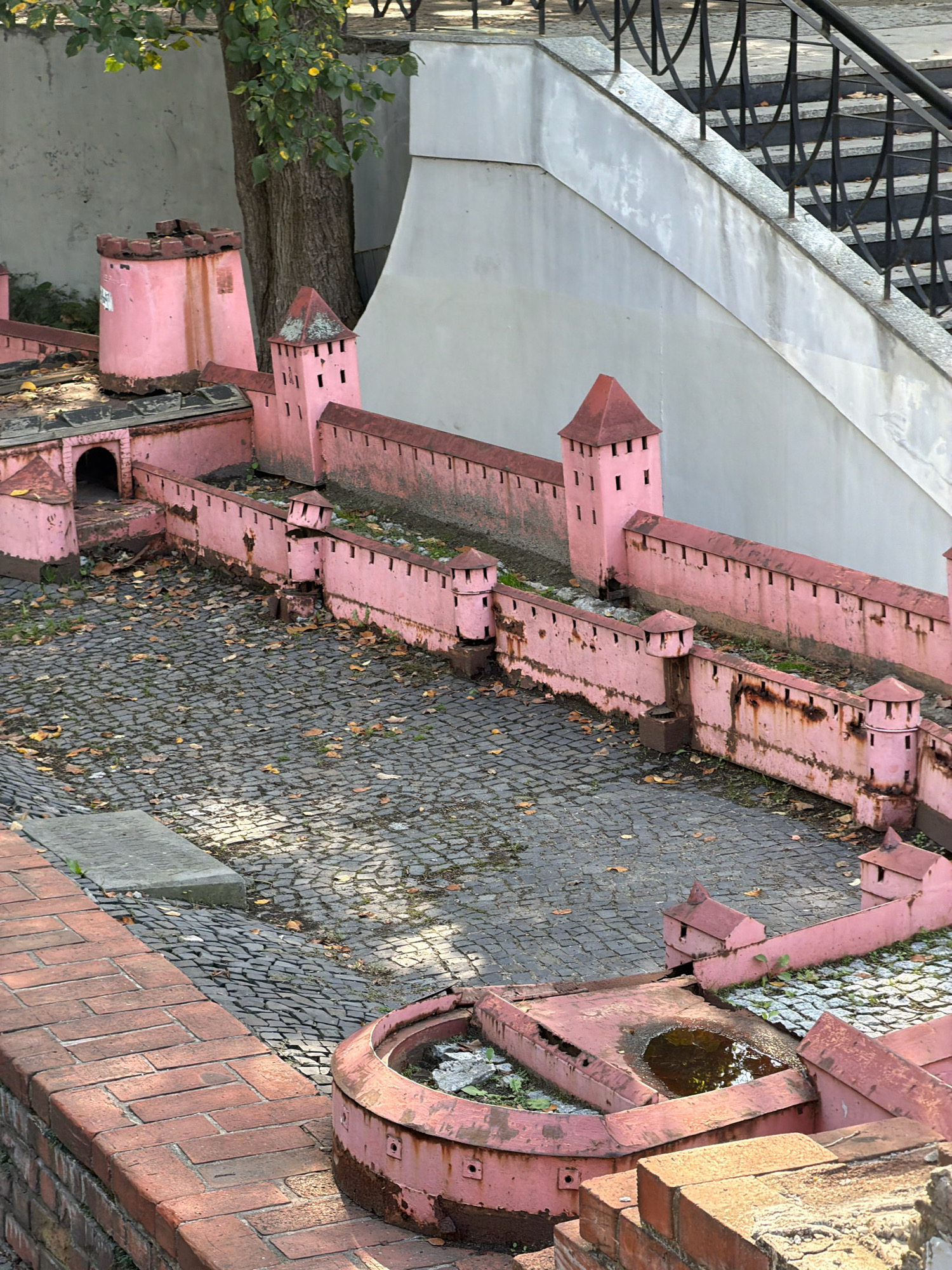 The image shows a miniature replica of a fortress, likely part of a larger exhibit. The fortress is painted pink and features walls, towers, and a gateway. The ground inside the fortress is paved with cobblestones, and scattered leaves suggest it is autumn. A tree with green and yellow leaves is visible behind the fortress. In the background, a concrete wall and stairs with a metal railing are present. The replica appears to be located in an outdoor urban setting, possibly a park or an open-air museum. A closer examination of the context and architectural style may indicate its specific location within Wrocław, Poland.