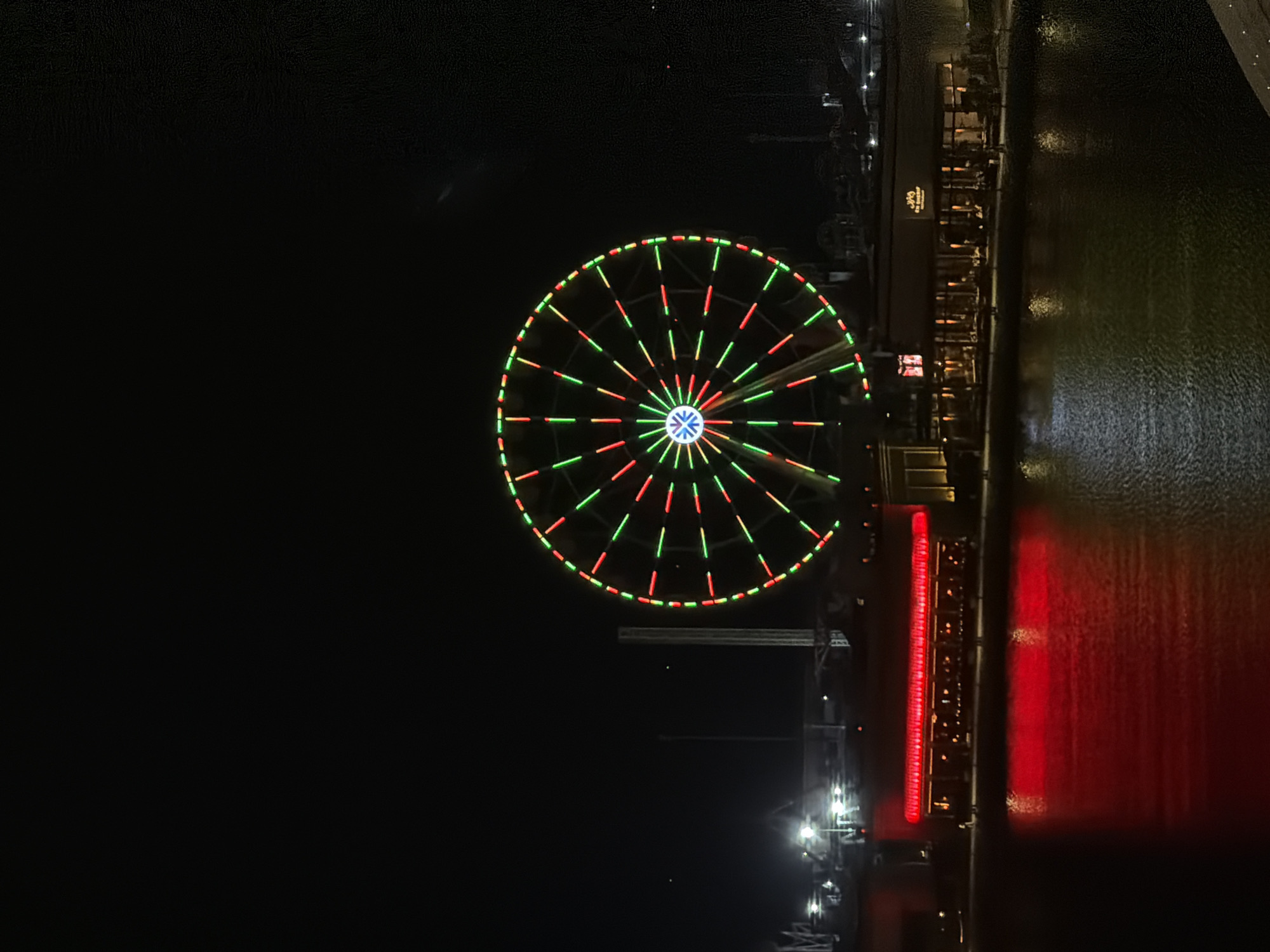 A nighttime scene features a large Ferris wheel illuminated with red and green lights against a dark sky. In the foreground, a body of water reflects the lights from buildings on the opposite shore. A long, red-lit building is visible on the left, and a building with a lighted sign is on the right. The environment appears to be an urban waterfront or entertainment district.