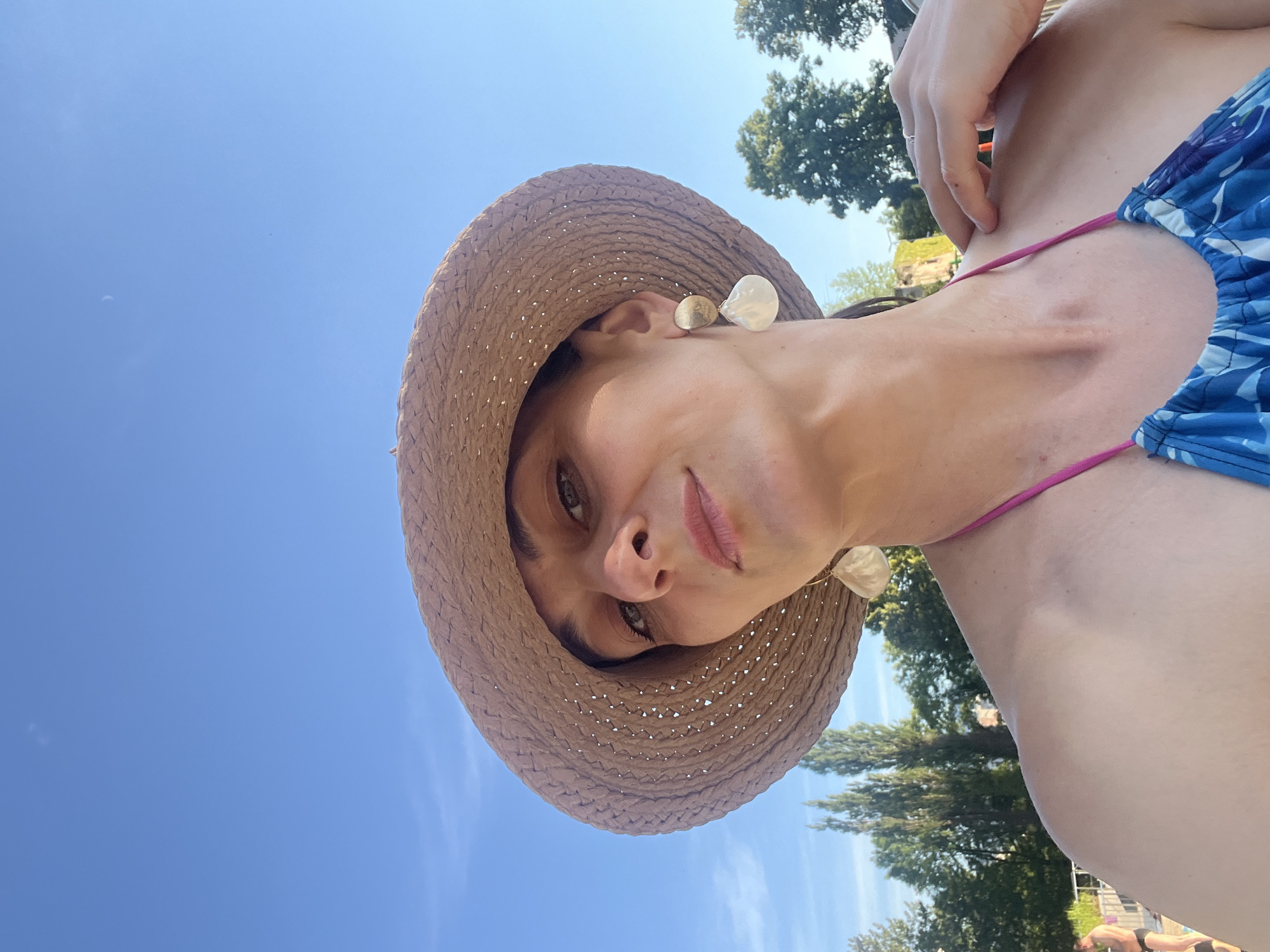 A woman is taking a selfie outdoors with a bright blue sky above. She is wearing a straw hat and a bikini top with a blue and white floral pattern. She has large, white shell-shaped earrings. The background shows green trees and a few people in swimwear in the distance. The woman's left shoulder is visible, and her right hand is resting on her right shoulder.