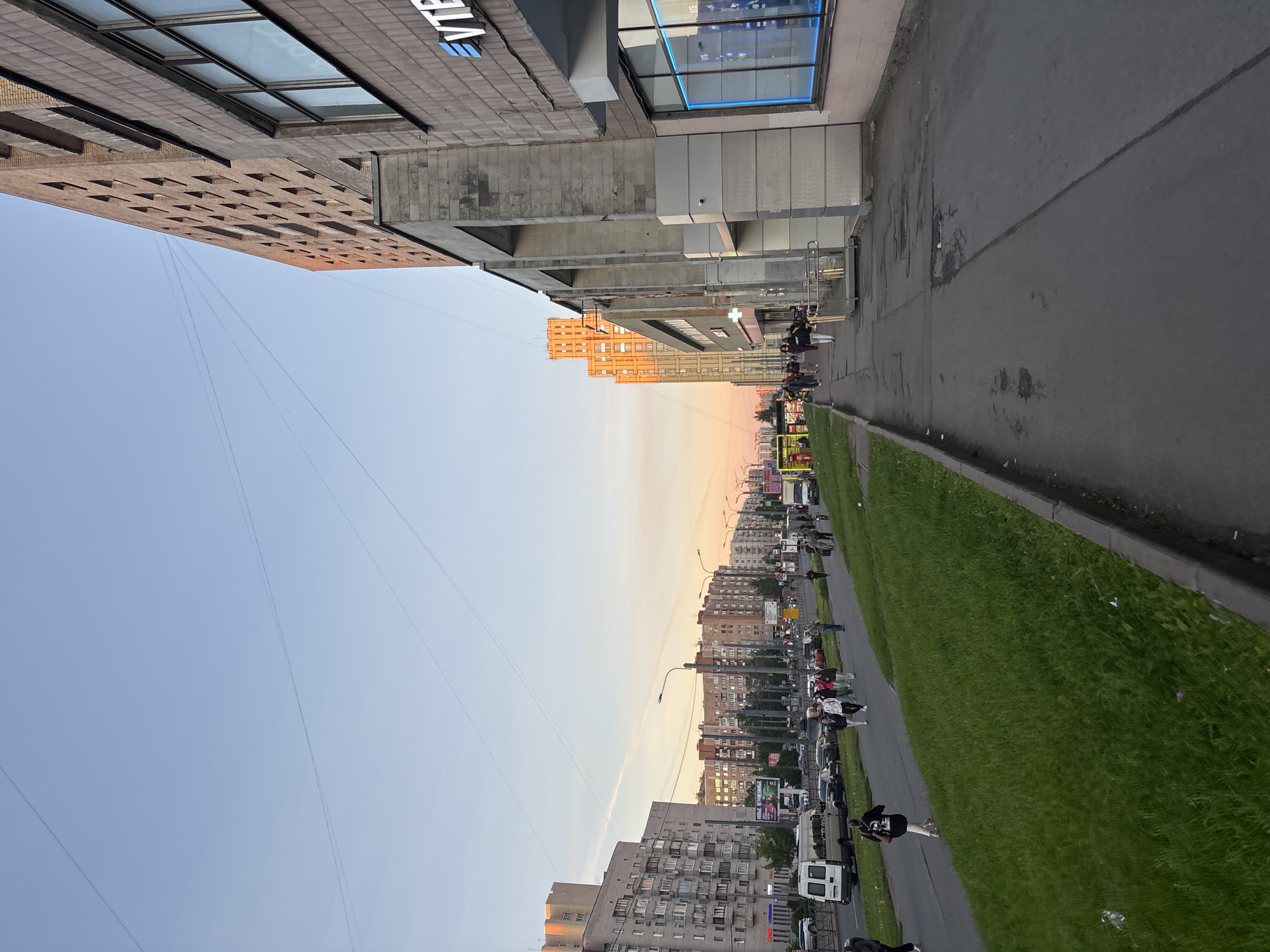 The image depicts a street scene in Sinyavino, Russia, likely during the late afternoon or early evening, as indicated by the warm, soft lighting and the clear sky with a hint of pink near the horizon. The perspective is from a sidewalk looking down a street lined with multi-story residential and commercial buildings.
On the left side of the street, there is a grassy embankment sloping down to a road with several lanes of traffic. Cars, including a white van, are visible on the road. A sidewalk runs parallel to the road, and several pedestrians are walking along it. Some are walking individually, while others are in small groups. The buildings on the left are residential, with numerous windows and balconies.
On the right side of the street is a modern commercial building with a facade of stone and glass. The building has large windows, some of which appear to be storefronts or offices. A prominent sign with blue lettering, "VTB", is visible on the building. The architecture suggests a relatively new development.
The sky is a clear, pale blue, with thin, almost invisible wires stretching across it, likely from utility poles. The overall impression is of a typical urban environment with everyday activity.
