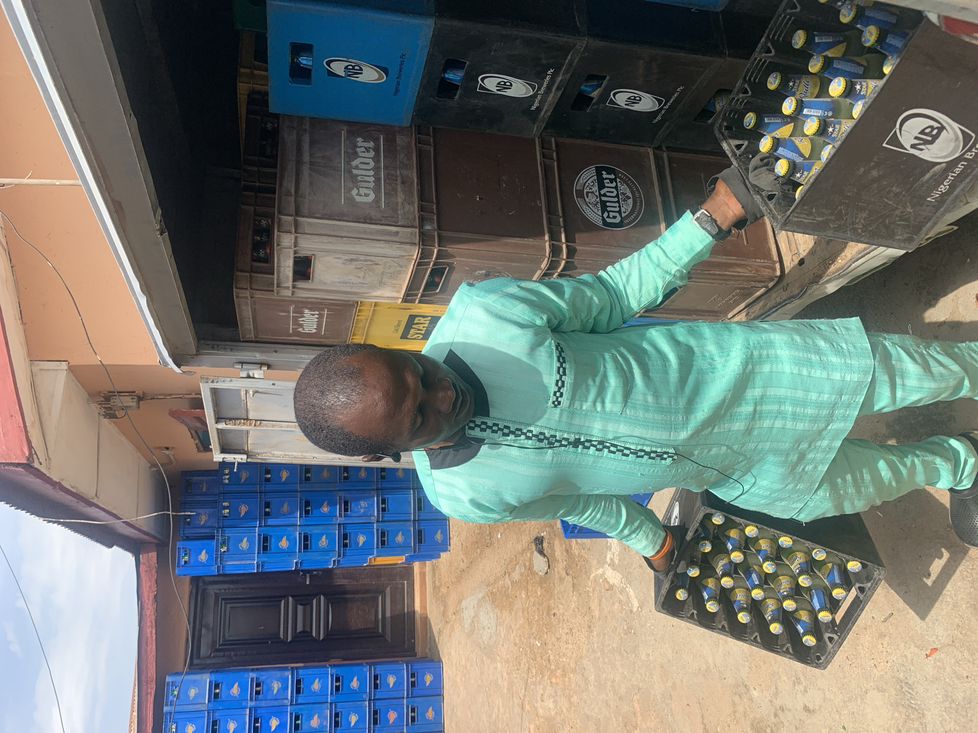 This image, set in what appears to be a commercial area in Jos, Nigeria, captures a man actively engaged in handling crates of bottled beverages under bright daylight. The man, possibly middle-aged, is seen wearing a light turquoise traditional tunic and trousers, featuring distinctive black and white patterned trim around the neckline and chest. He has a black watch on his left wrist and wears dark shoes.
He is in the process of loading or unloading crates, holding one black plastic crate full of bottles at waist height and another, seemingly empty or partially full, lower down. The bottles predominantly feature yellow and blue labels with yellow caps, identifiable as "STAR" and "Radler" brands.
The background is filled with numerous stacks of plastic crates. Directly behind the man, brown and black crates are visible, bearing the "Gulder" logo and "NB Nigerian Breweries Plc." Some blue crates also display "NB Nigerian Breweries Plc." A yellow crate, marked "STAR" and "Cold Filtered," is partially visible. To the left, more blue crates are stacked against a building with a tan-orange facade and a dark wooden door. The clear sky and strong shadows indicate a sunny day. The scene depicts a common sight in beverage distribution or retail in the region. Visible text includes "Gulder," "STAR," "NB Nigerian Breweries Plc," "Radler," "Cold Filtered," and "International Quality Beer" (on a Gulder crate).
