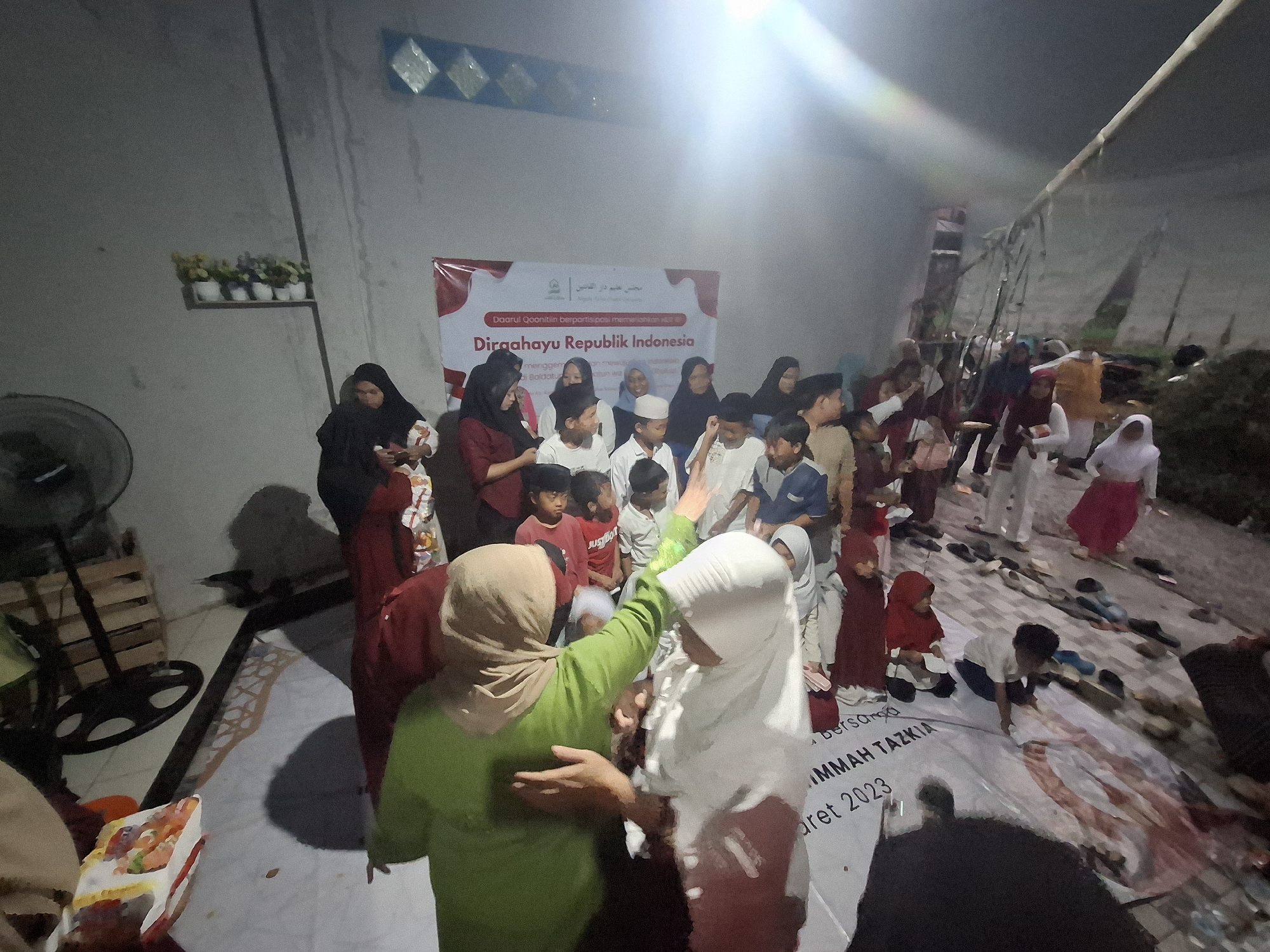 In Babakan Madang, Indonesia, a group of approximately 30 people, predominantly children and women, are gathered indoors. The event appears to be a celebration of Indonesian Independence Day, as indicated by a banner in the background that reads "Dirgahayu Republik Indonesia". The banner also includes Arabic text and the logo of "Darul Qonitin Berpartisipasi".
The individuals are dressed in a variety of attire, with many women wearing headscarves and modest clothing. The children are in casual wear, some in t-shirts and shorts, others in more formal outfits. Several children are wearing traditional Indonesian peci caps.
The environment is dimly lit, suggesting it is either evening or a poorly illuminated indoor space. The walls are a plain, textured gray. A shelf with several small potted plants is mounted on the wall. A large, industrial fan is visible on the left side of the image.
On the floor, a large banner is spread out, displaying text related to "JIMAH TAZKIA" and " Maret 2023". Several pairs of shoes and sandals are neatly placed on the floor around the gathering. A few individuals are interacting, with some women appearing to guide or assist the children. The overall atmosphere seems to be one of community and celebration.