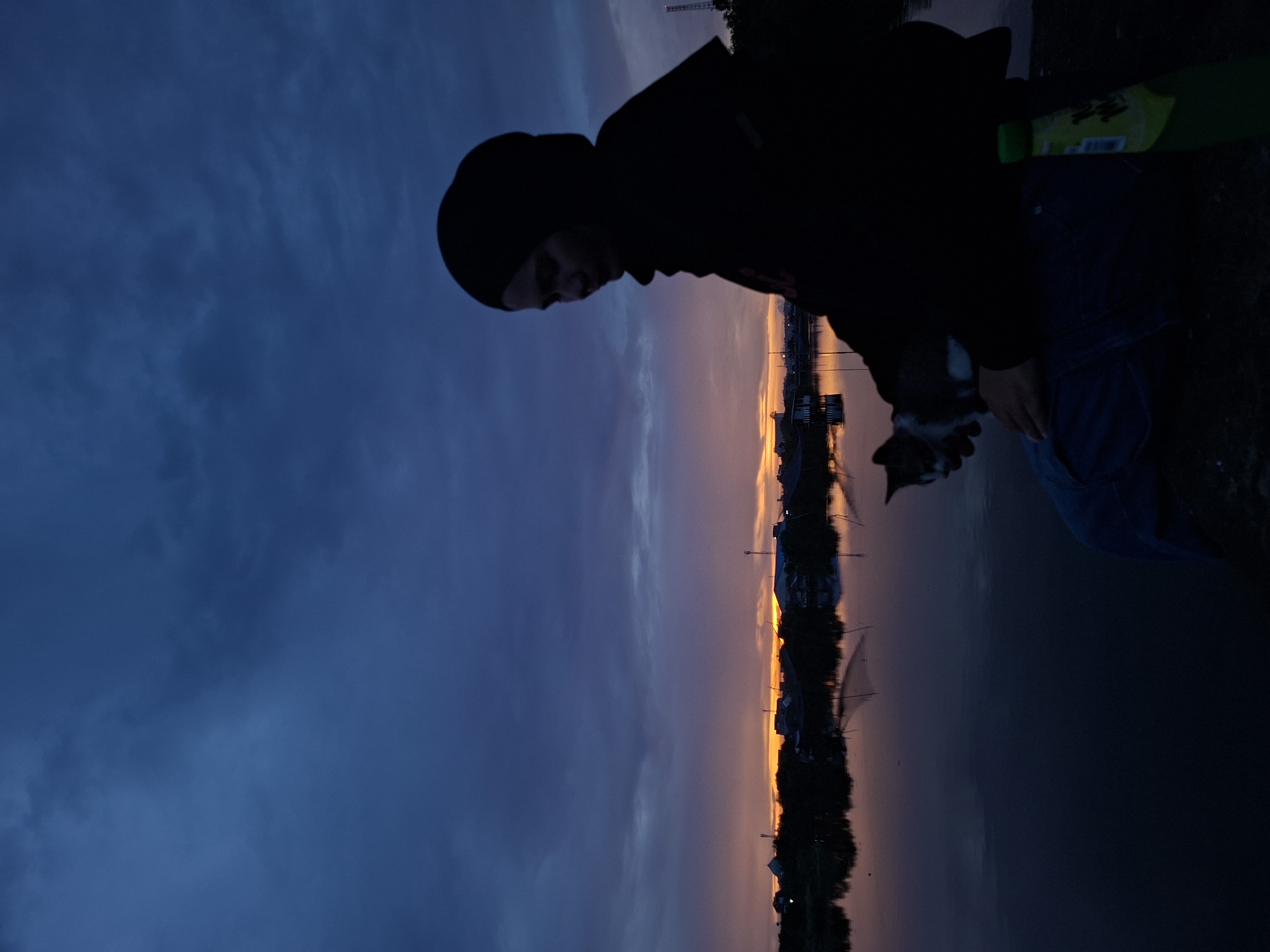 A person wearing a black beanie and dark clothing is holding a cat at sunset near the city of Lhokseumawe, Indonesia. The sky is a mix of dark blue and orange, reflected in a body of water below. Buildings and trees are silhouetted against the sunset. The person appears to be standing on a grassy area.