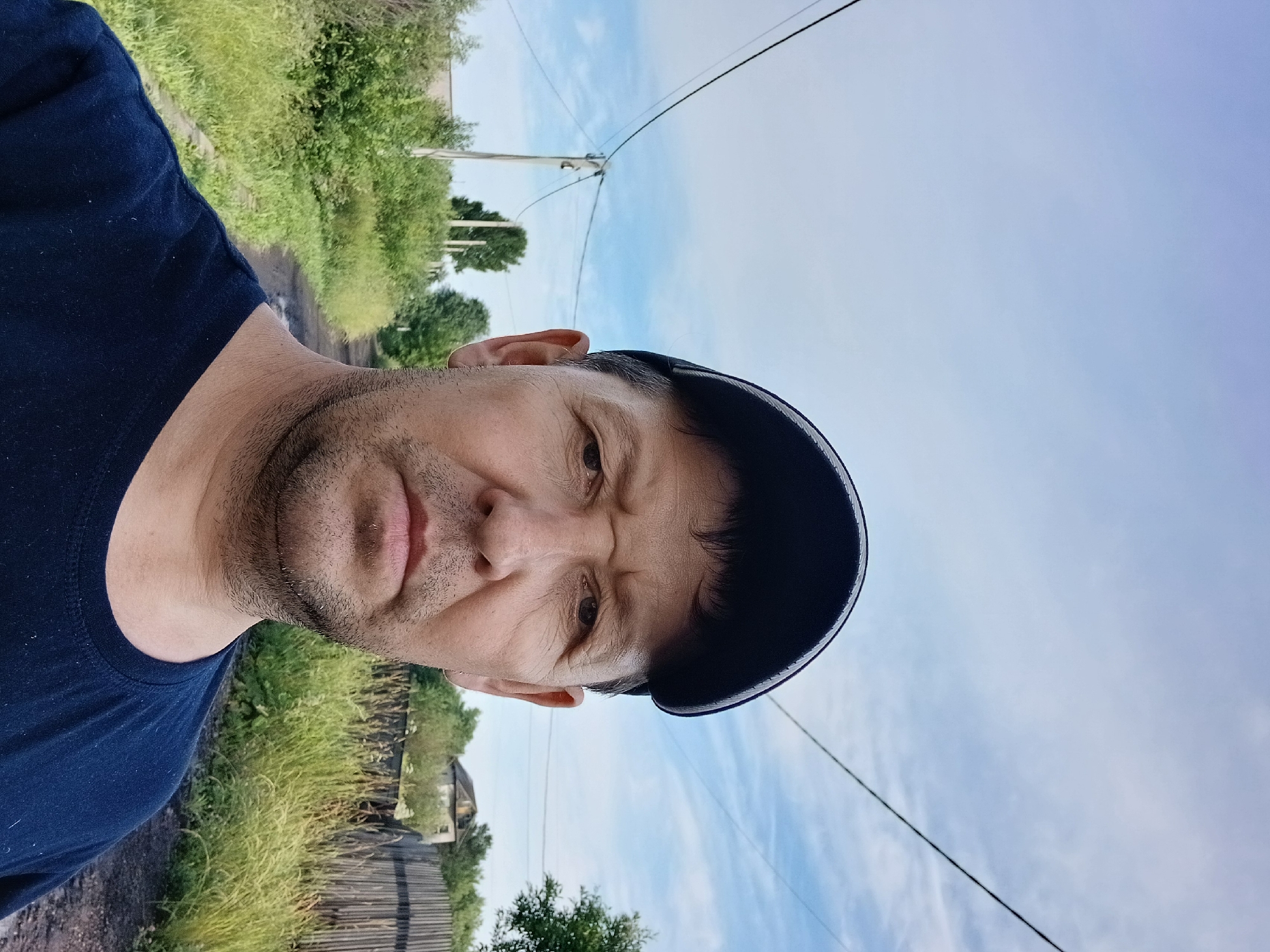A man wearing a dark blue t-shirt and a black baseball cap is photographed in a close-up selfie. He has a light complexion and short, dark hair visible beneath the cap. His expression is neutral. The background shows a rural or semi-rural setting, possibly in Prokopyevsk, Russia. A dirt road, flanked by overgrown grass and vegetation, recedes into the distance. Power lines are visible crisscrossing the sky, supported by a utility pole. A wooden fence and the roof of a house are discernable further down the road. The sky is partly cloudy with a mix of blue and white. The lighting suggests it is daytime, possibly late afternoon or early morning due to the soft light.