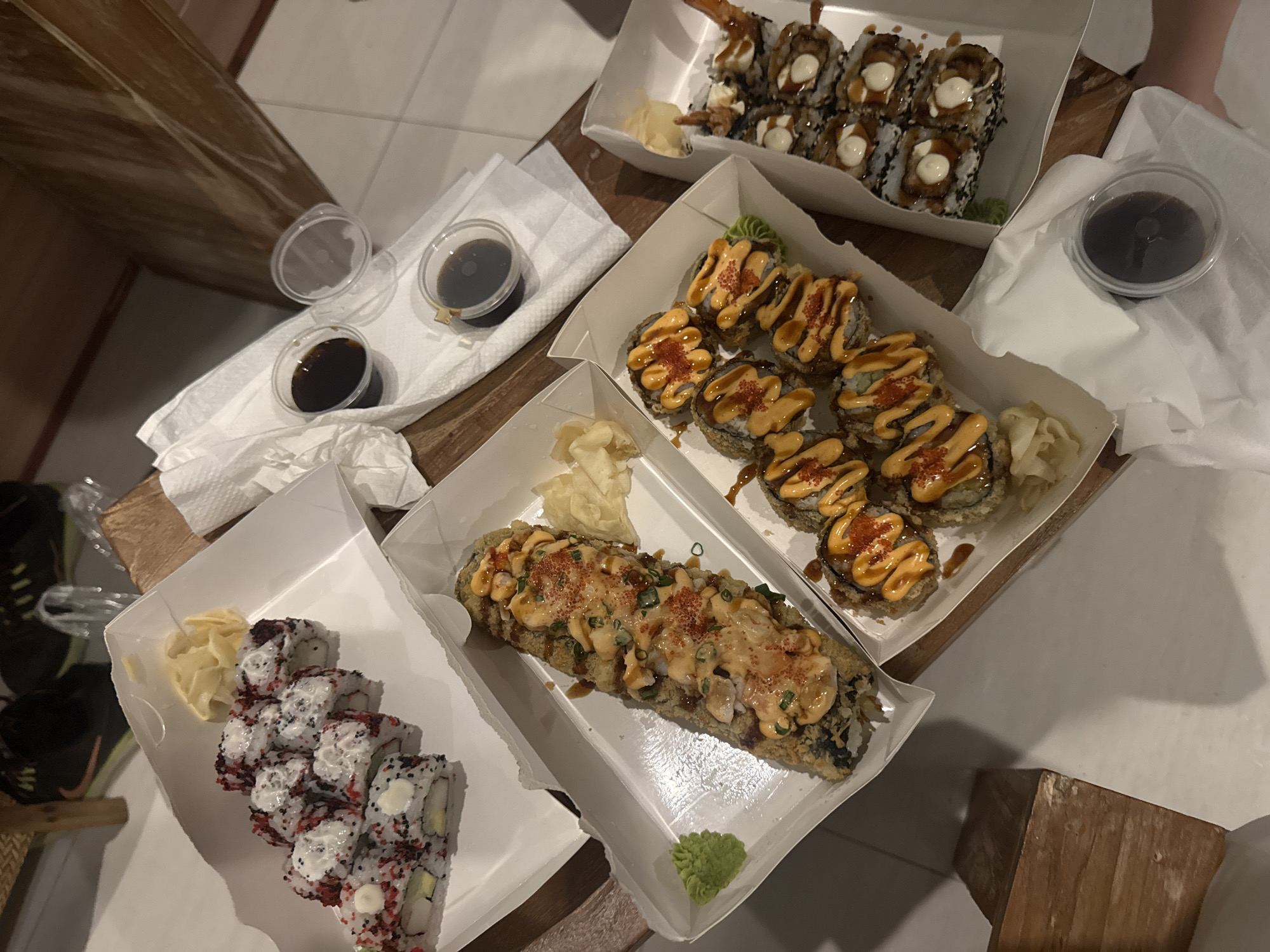 This image captures a spread of sushi takeaway meals laid out on a light-colored wooden table. The scene presents a casual dining experience, likely indoors at a home or hotel room, given the presence of a white tiled floor and glimpses of other personal items like shoes in the background.
There are five white cardboard takeaway containers, each holding different types of sushi. The container at the top features sushi rolls with a dark, possibly black sesame, coating, topped with a creamy white sauce and what appears to be fried shrimp or tempura. The middle container holds several round sushi rolls that are deep-fried or tempura-battered, generously drizzled with a creamy, orange-red spicy sauce and sprinkled with red specks, possibly tobiko. A dollop of green wasabi and pickled ginger are also present. The lower-middle container showcases a single, elongated, fried sushi roll, similarly topped with the orange-red sauce and red specks, accompanied by wasabi and ginger. In the lower-left container, sushi rolls with a white rice exterior are visible, speckled with red and black toppings and containing a creamy white filling.
Numerous small, clear plastic cups of dark soy sauce are placed on white paper napkins around the sushi containers. Pickled ginger is consistently provided in most of the boxes. The lighting appears to be artificial, suggesting an evening or night time. No people are visible, and there is no discernible text on any of the packaging. The overall arrangement suggests a meal ready for consumption.