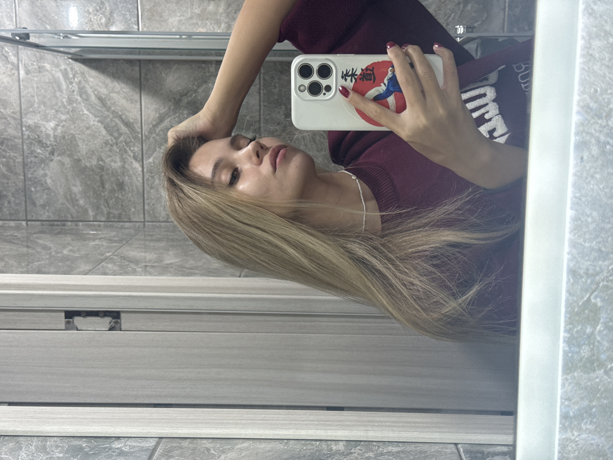This media file is a vertically oriented, mirror selfie of a young woman. She has long, blonde hair that is styled over her shoulder. She is wearing a maroon sweater with white text on the sleeve that reads "BUD". In her right hand, she is holding a white smartphone, also positioned to take the photo. The phone case features a circular design with Japanese characters and an illustration of a person. Her nails are painted a deep red.
The woman appears to be in a bathroom, suggested by the tiled walls and what looks like a shower enclosure. The lighting is soft and diffuse, not indicating a specific time of day. Her expression is relaxed, with her eyes closed and a slight smile on her lips. The overall mood is casual and personal. The location context of Chaikovsky, Russia, is not directly discernible from the visual elements, but the image itself captures a private moment.