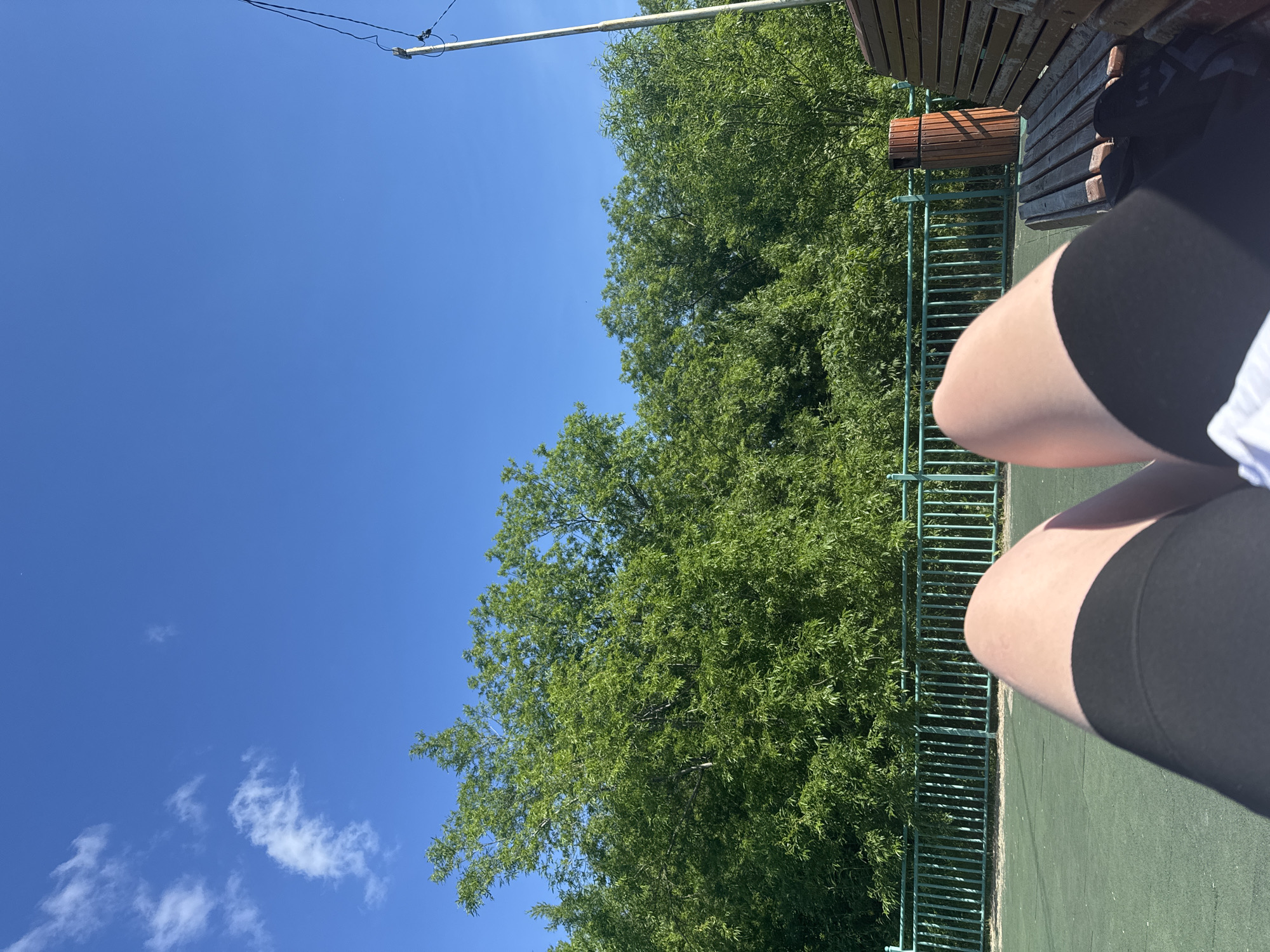The image depicts a sunny day with a clear blue sky. In the foreground, a person's legs are visible, wearing black shorts. The person appears to be sitting on a park bench made of wood, with some black material visible on the bench. To the left of the bench, there is a green fence and lush green trees in the background. A wooden trash bin is situated next to the bench. A utility pole with wires is visible on the right side of the image. The ground appears to be a green, possibly rubberized surface, common in outdoor recreational areas. The scene suggests a casual outdoor setting, likely a park or a recreational facility within the city of Petropavlovsk-Kamchatsky, Russia.