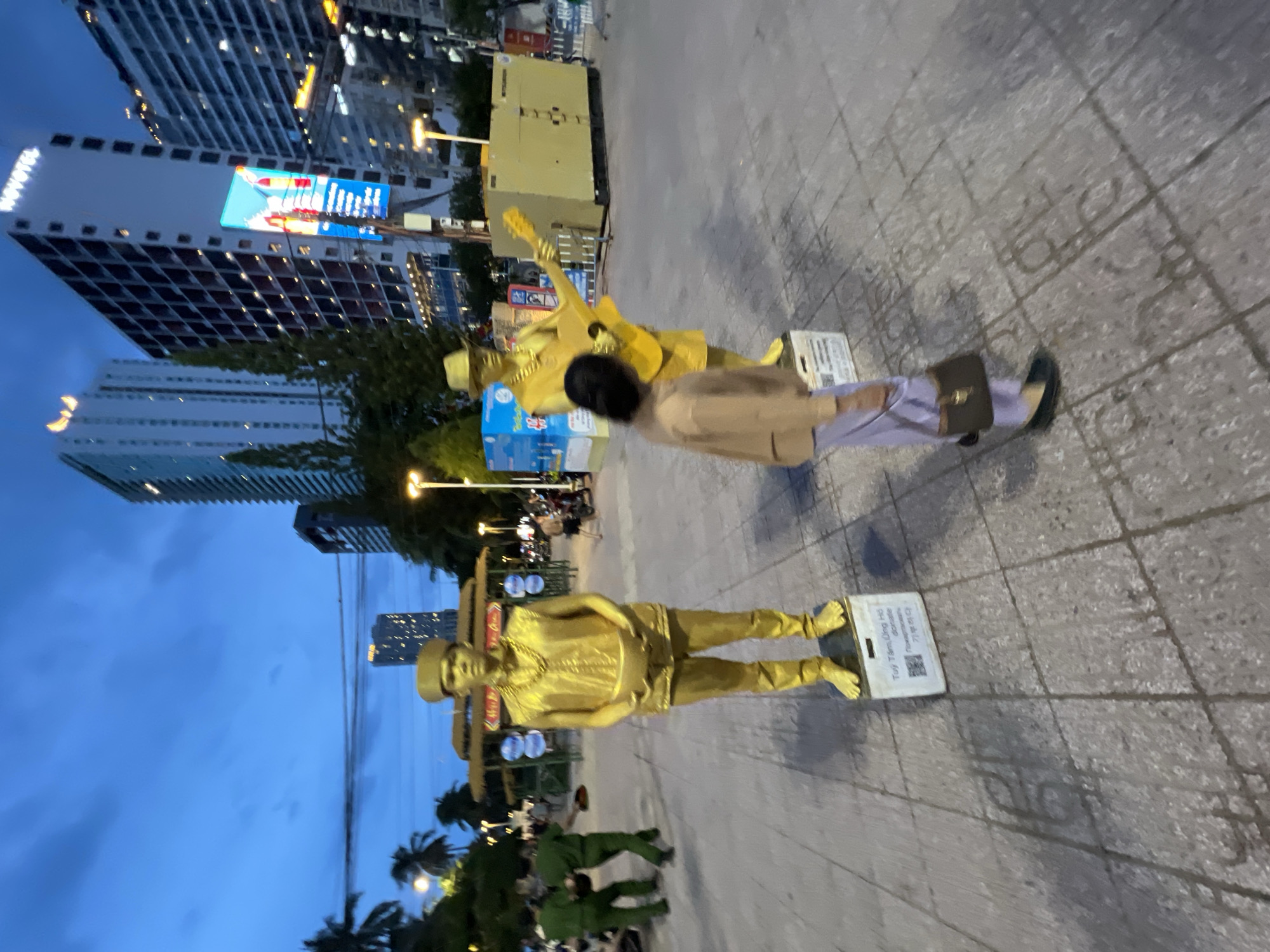 The image depicts a public square in Nha Trang, Vietnam, at dusk. In the foreground, a person dressed in a light brown long-sleeved shirt and light purple pants, carrying a dark brown handbag, walks away from the viewer towards the right. To the left, two individuals in green uniforms, possibly security personnel, are walking towards the left.
The main subjects are two street performers covered in gold paint. The performer on the left stands on a white base, holding a bowl. They are wearing a gold vest, gold pants, and a gold necklace, with a gold cap on their head. The performer on the right, also standing on a base, is playing a gold guitar and wearing a gold hat and a gold shirt and pants.
In the background, several tall buildings are visible, including a Novotel hotel. There are also palm trees and streetlights. The sky is a deep blue, indicating early evening. The ground is paved with stone tiles. A large yellow generator-like structure is present on the right side of the image, with fencing behind it. Colorful banners and signs are also visible in the background.