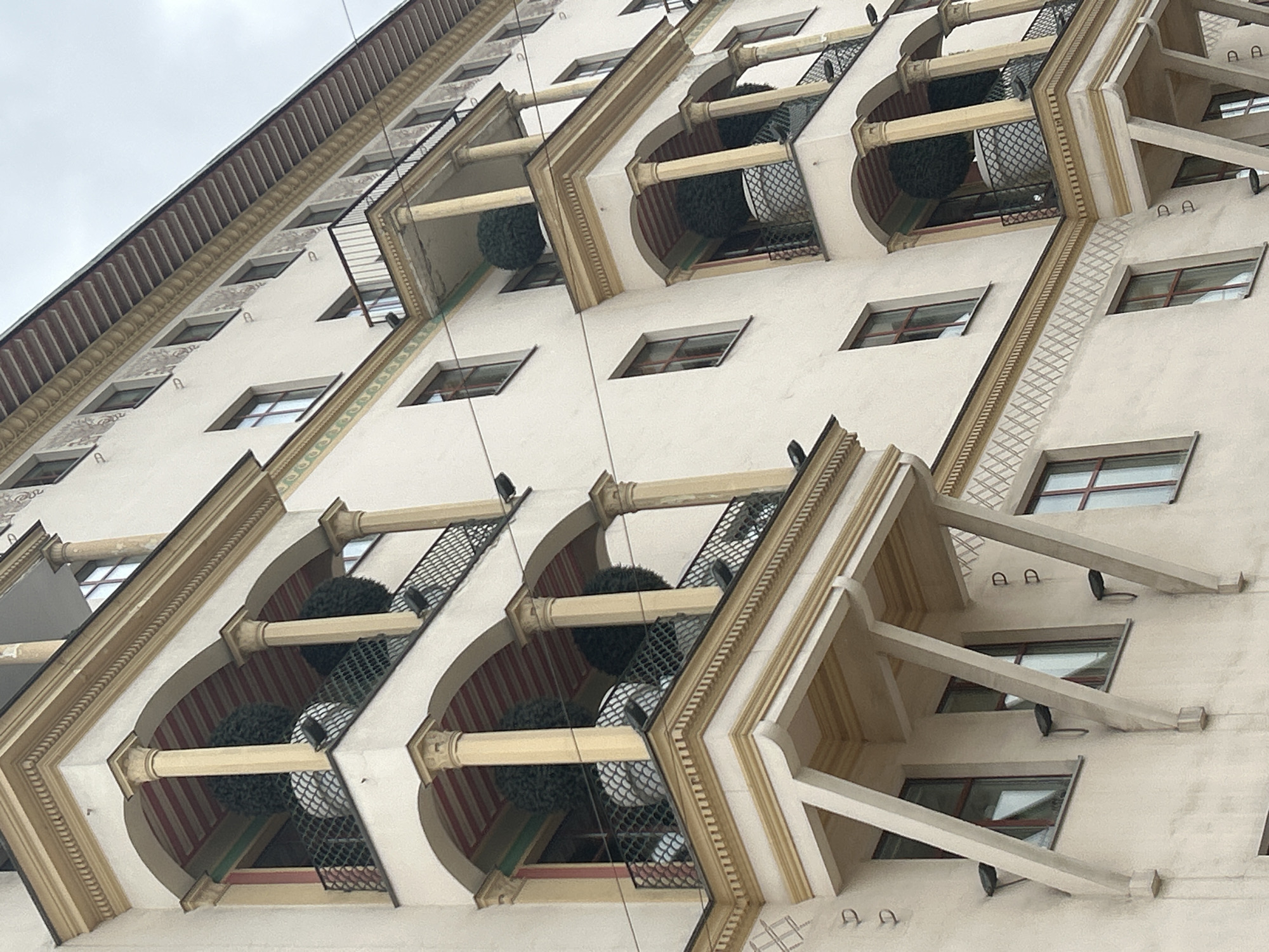 An upward-angled view captures the ornate facade of a building in Moscow, Russia, under an overcast sky. The multi-story structure features a light beige stucco exterior adorned with elaborate architectural details.
The building is characterized by a series of projecting bay windows or recessed balconies, which are distinctively arch-shaped. Each arch is framed by decorative gold-toned trim and supported by small columns with capitals. Within these arched recesses, large, dark green spherical objects, resembling topiary plants, are visible. A black mesh or netting covers parts of the opening, and some interior walls of these spaces are decorated with red and white vertical stripes.
Between these prominent arched features and on higher levels, the facade contains numerous rectangular windows with dark frames. Horizontal bands of intricate, geometric patterns in gold, brown, and hints of green run across the building, adding to its decorative appeal. The uppermost section features a dark, ribbed cornice or roofline. On the plain wall sections, particularly visible in the crops, small, stylized black "A" shapes appear as a repeating decorative motif. On the right side of the image, angular, light-colored concrete supports extend from the building's structure.
No people or specific activities are visible, emphasizing the architectural grandeur of the building. The overall impression is one of a richly detailed and well-maintained historic or classical revival building.