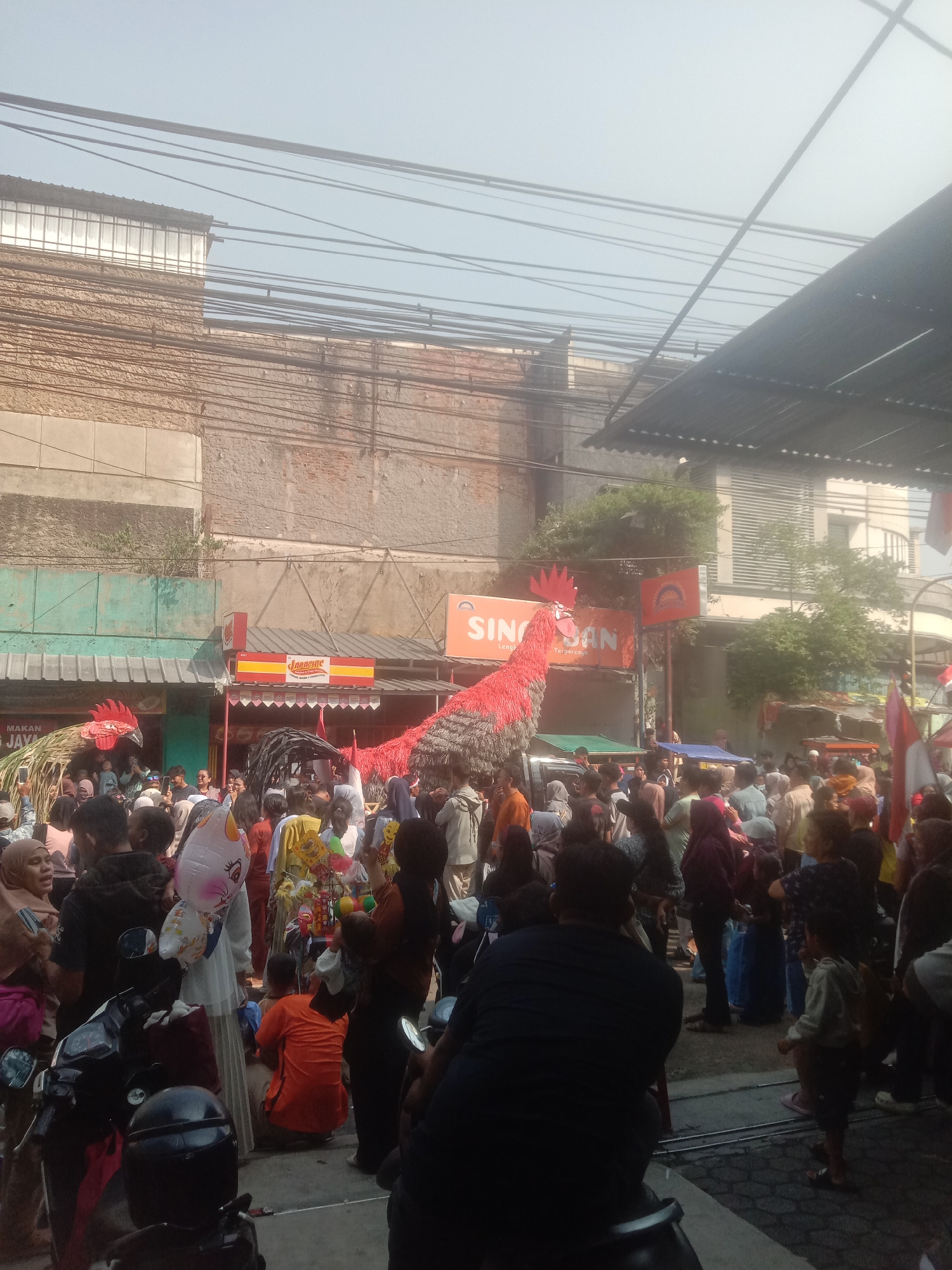 The image depicts a street scene in Cianjur, Indonesia, likely during a public gathering or parade. In the foreground, several motorbikes are parked. A crowd of people, dressed in casual attire, are gathered, many looking towards the center of the image. Dominating the background are two large, red, rooster-shaped effigies. One rooster effigy is more fully visible on the left, constructed with straw-like material for its body and a red, feathery head. The other rooster effigy is larger and more central, appearing to be made of fabric and possibly wire, with a prominent red comb and tail feathers. Businesses with awnings and signs are visible behind the crowd, one sign reads "SIN SAN Lengkap & Terpercaya" which translates to "SIN SAN Complete & Trusted". The architecture suggests a commercial or residential area with brick buildings and utility wires overhead. The sky is hazy and bright, indicating daylight. Some individuals are holding up balloons and flags, contributing to the festive atmosphere. People are engaged in various activities, including observing the rooster effigies, conversing, and moving through the crowd. The overall scene conveys a sense of community engagement and cultural celebration.