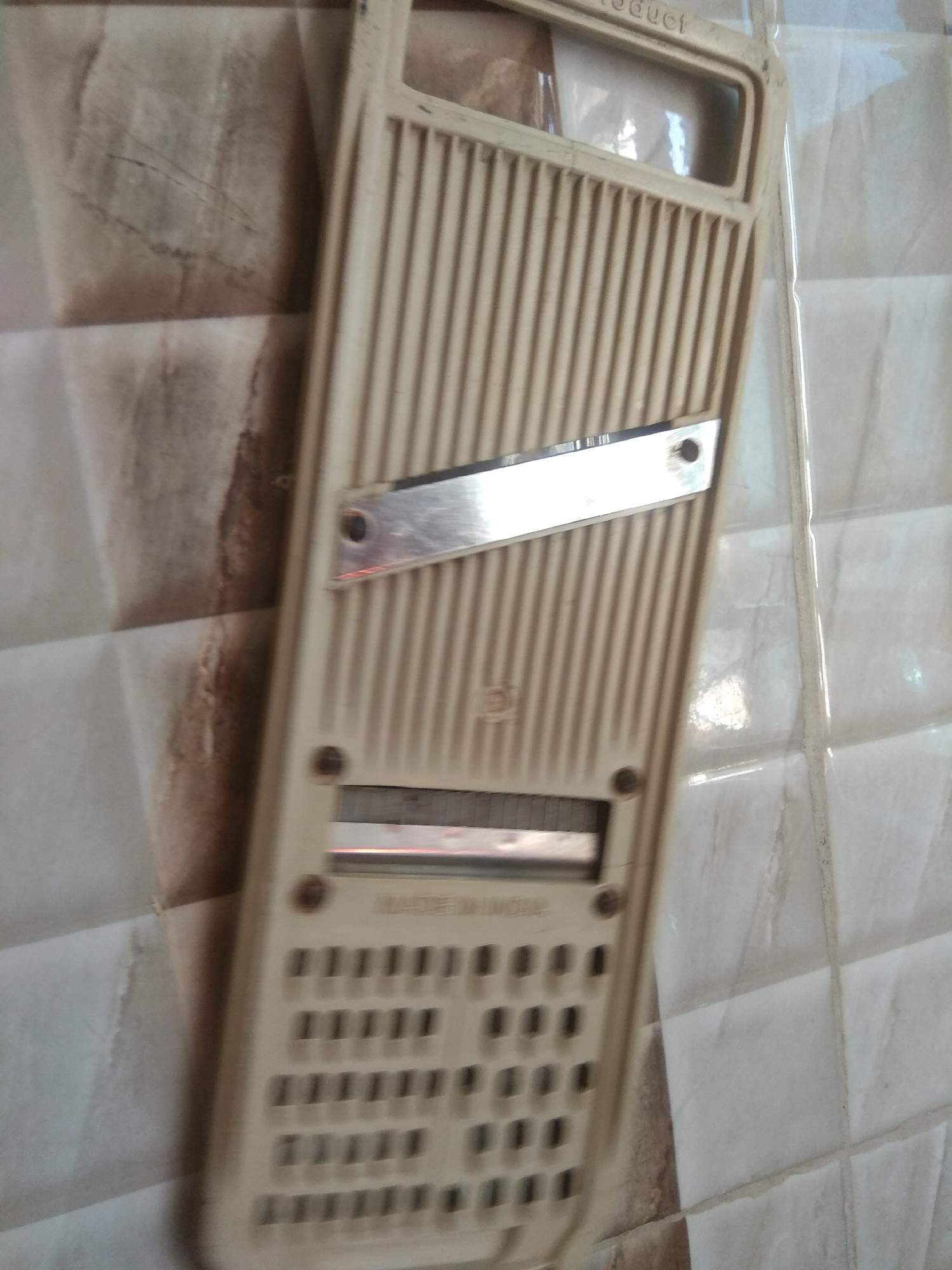 The image shows a close-up, slightly angled view of a beige manual grater or mandoline slicer. The grater is positioned against a tiled wall with a marble-like pattern in shades of beige and brown. The wall tiles are square and have a slightly raised, beveled edge, creating a subtle grid effect.
The grater itself has a textured surface with parallel vertical ridges. A shiny metal blade is attached diagonally across the upper portion of the grater, secured by two small screws. Below the blade, there is a slot with another metal component visible, likely a cutting surface or interchangeable blade. Further down, there are rows of small, rectangular openings, suggesting different grating or slicing options. The grater appears to be made of plastic and has a utilitarian design.
At the very top of the grater, a faint inscription is visible. The letters are uppercase and appear to read "PRODUCT". The image is well-lit, with soft, diffused light that highlights the textures of both the grater and the wall. There are no people or animals visible in the image. The setting appears to be a domestic kitchen or food preparation area. The time of day and weather conditions are not discernible from the image. The overall impression is one of a functional kitchen tool against a neutral background.