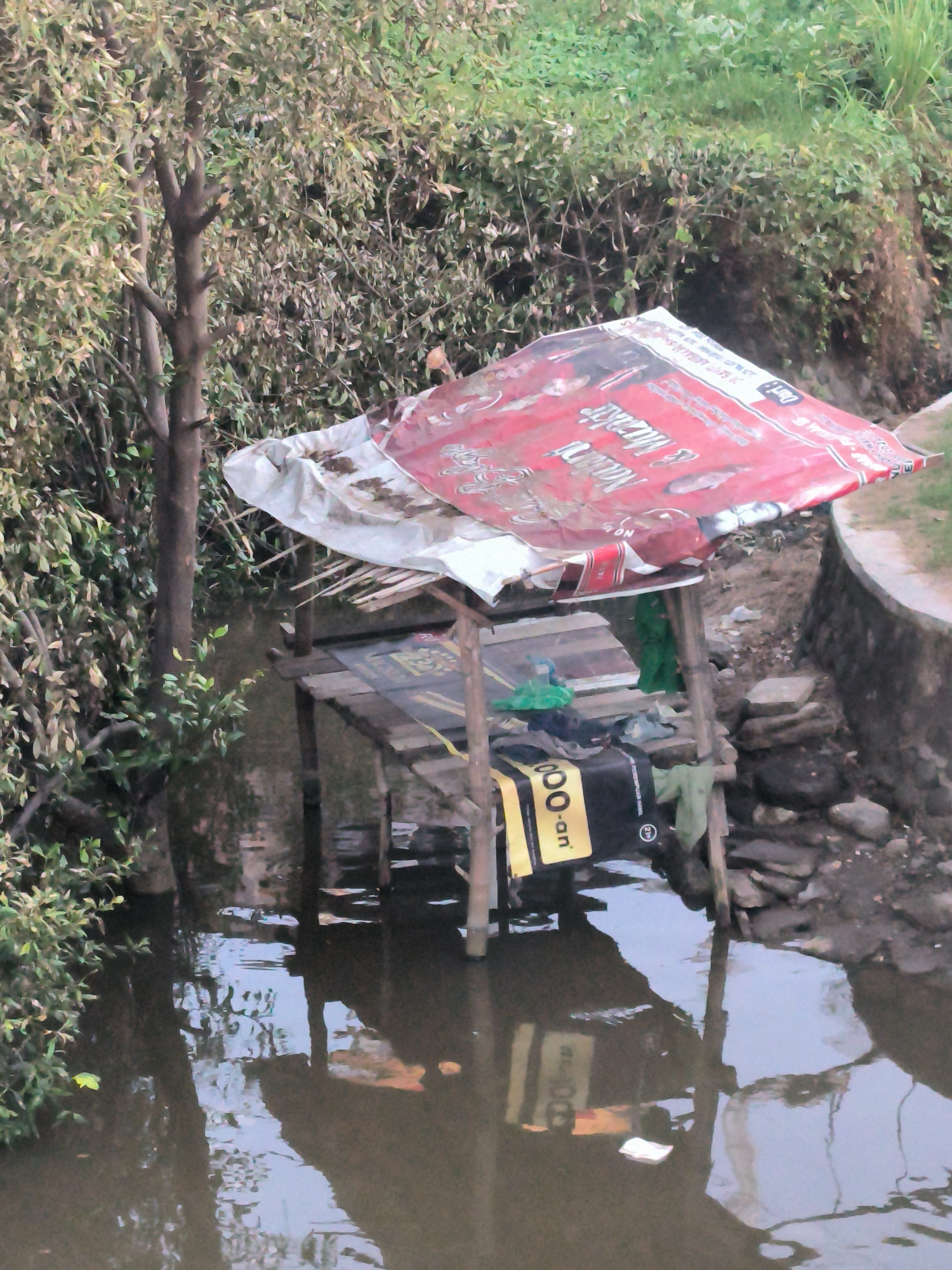 A ramshackle shelter built over a body of water is constructed of wooden beams and covered with a damaged promotional banner. The water reflects the structure and the litter and debris scattered around it. The shelter is located beside a narrow concrete path along the water's edge, in an overgrown area of foliage. The location is Lhokseumawe, Indonesia.