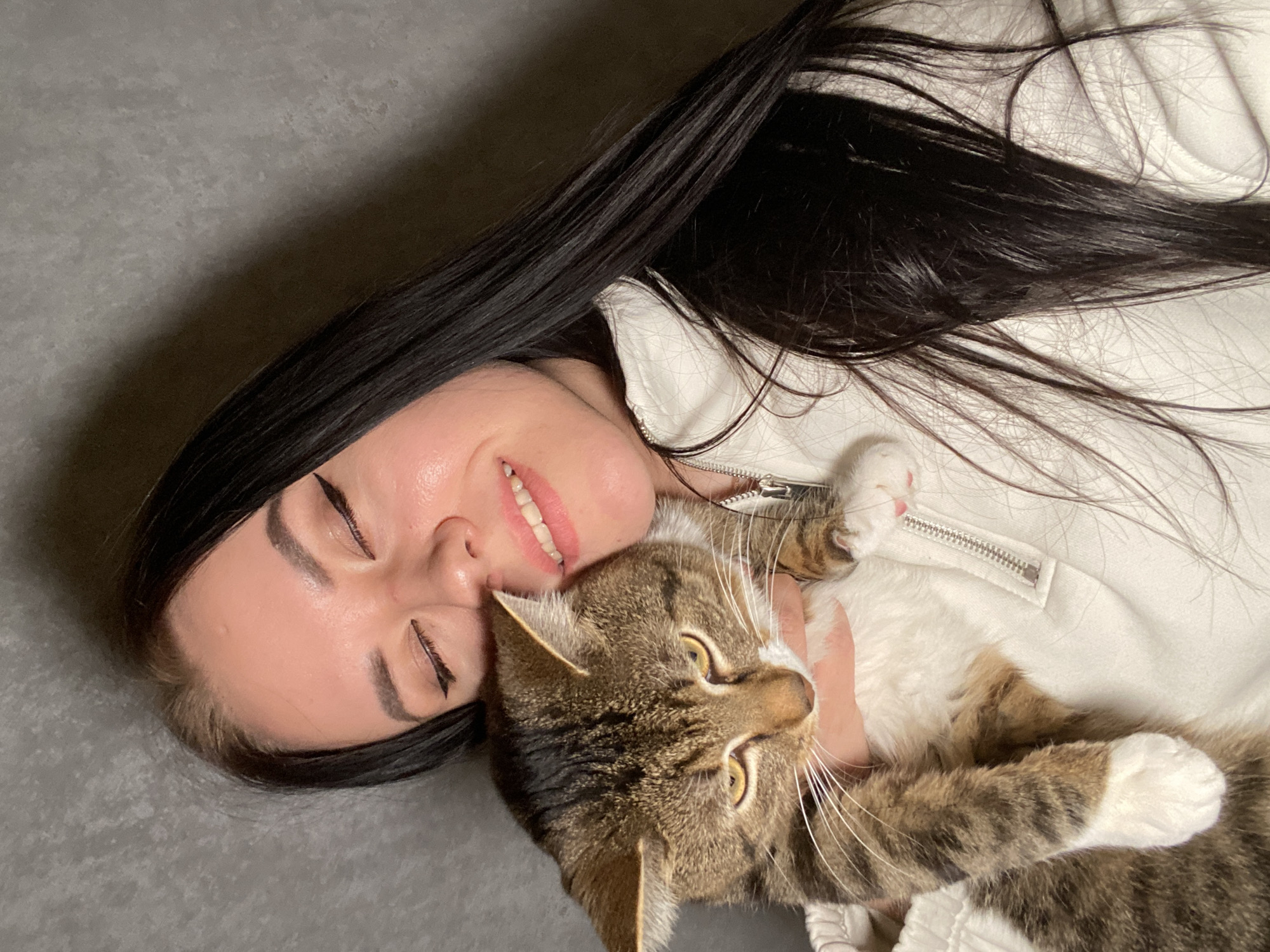 A woman with long black hair lies on a grey surface, smiling slightly with her eyes closed. She wears a white zippered jacket. A brown and white tabby cat lies on her chest, with its head facing the camera. The location is unknown and cannot be determined from the image.
