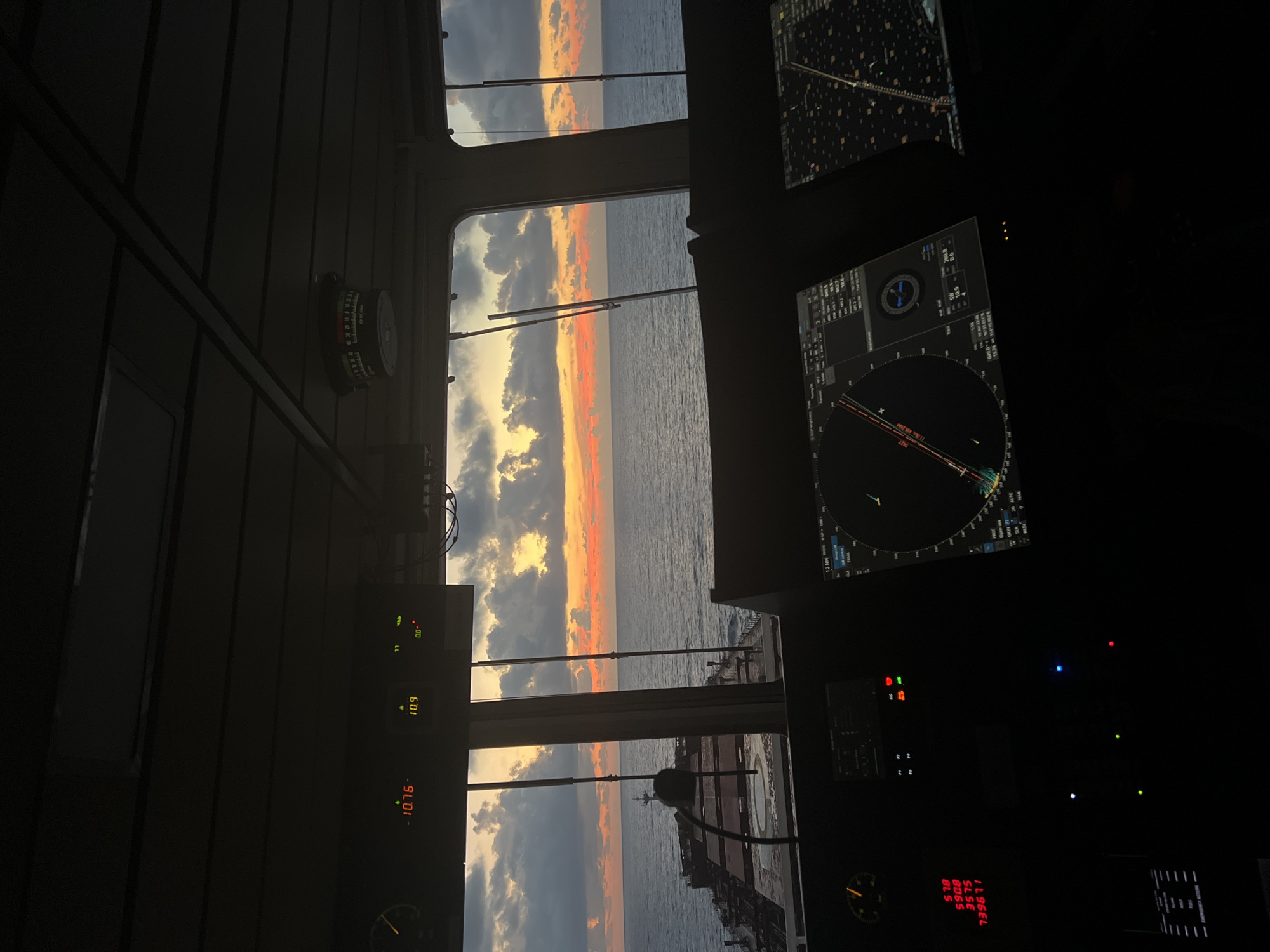 The image shows the interior of a ship's bridge at sunset. Through the large windows, the ocean and a dramatic sky with clouds are visible. A large cargo ship is seen in the distance, partially obscuring the horizon. Inside the bridge, several electronic displays and instruments are present, including radar screens and various readouts with numbers and symbols. The lighting is dim, with the glow from the screens and the sunset providing illumination. The overall scene suggests a setting at sea during twilight.
