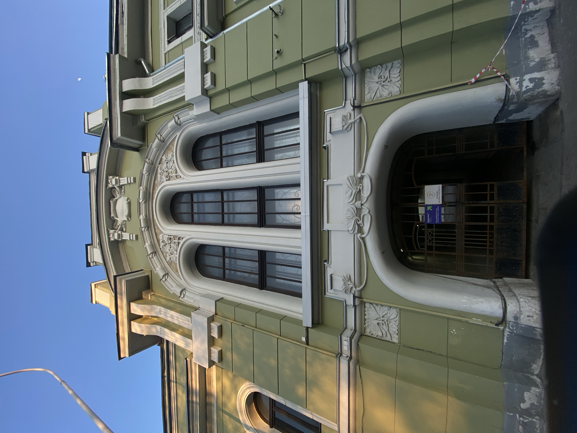 The image depicts the facade of an Art Nouveau building located at Bolshoy Kozikhinsky Pereulok, 13/27, in Moscow, Russia. The structure is painted pale green with white architectural trim, details, and decorative stucco work.
The ground level features a large arched entrance secured by an ornate dark metal gate, which is partially open on the right side. Signs attached to the gate identify the premises as "Ренессанс" (Renaissance) and "КЛИНИКА ДОКТОРА КРЕМНЕВА" (Doctor Kremnev's Clinic). A red-and-white striped caution tape is draped across the lower right section of the archway.
Above the entrance, three prominent arched windows with dark frames and multiple panes are centrally located on the upper floor, flanked by white pilasters and intricate floral or organic stucco relief work. A smaller, similar arched window is visible on the far left. The upper section of the building includes a decorative cornice and various architectural projections, some reflecting sunlight. Two small black security cameras are mounted on the green wall to the right of the central windows, and a silver downspout is visible on the far right.
The sky above is clear blue, with a faint crescent moon visible in the upper right. A street light arm extends into the frame from the upper left. Sunlight illuminates the upper facade, creating contrasts between light and shadow. No individuals or actions are depicted within the scene.