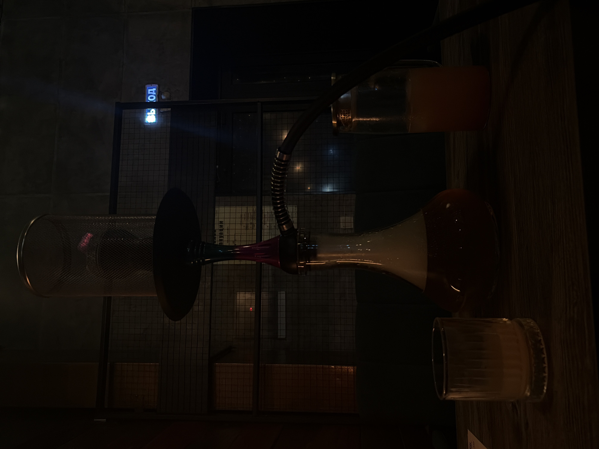 An indoor, dimly lit scene features a hookah pipe with a glass base filled with amber liquid and smoke. Next to it are two glasses, one containing a light-colored liquid and the other a darker amber liquid. A blue illuminated sign with Russian text "ВЫХОД" (Exit) is visible on the wall in the background. The setting appears to be a bar or lounge area, indicated by the seating and rustic wooden table.