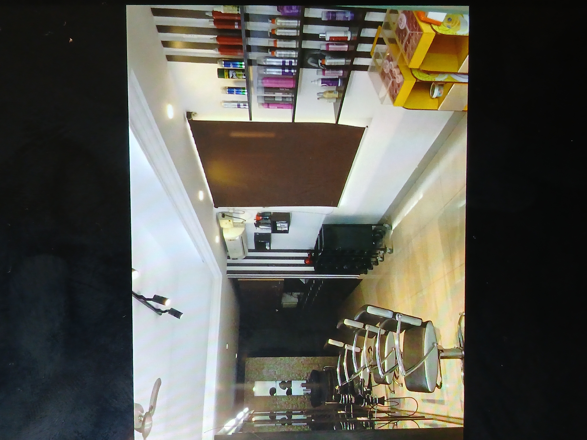 The image depicts the interior of a salon. It appears to be a modern or recently renovated space with recessed lighting and a brown rectangular accent panel on the ceiling. Shelves filled with various hair products line one wall, while yellow containers are stacked nearby. The salon features barber chairs, and black equipment is situated along another wall. A white air conditioning unit is mounted near the ceiling. The location is likely in a city, potentially Bengaluru, India, given the visual clues, though this cannot be definitively confirmed.