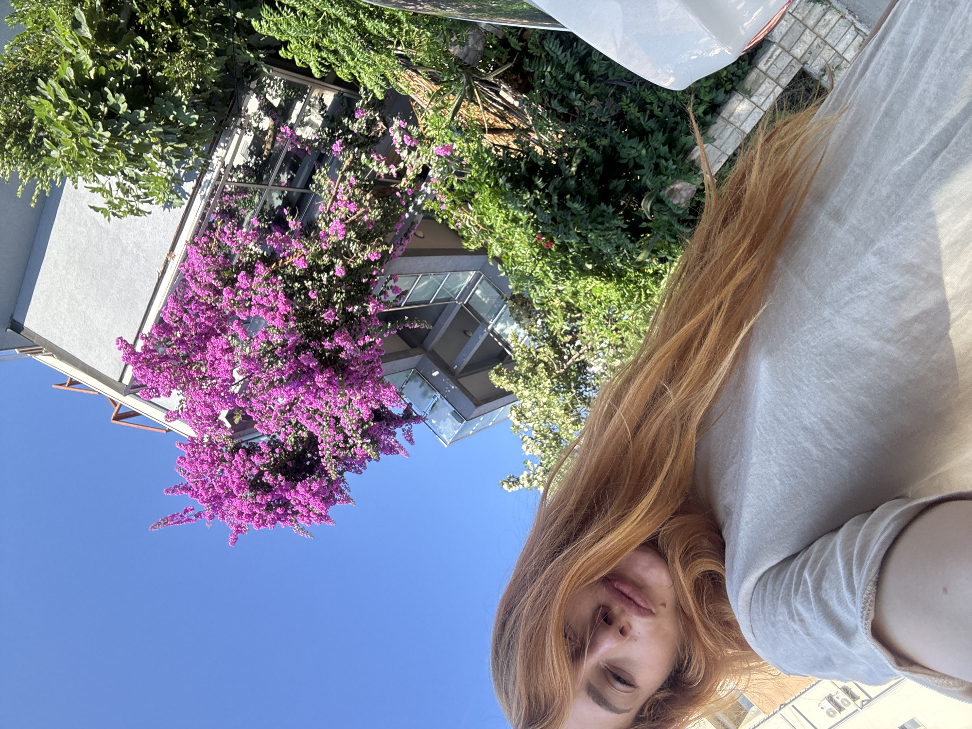A woman with long, auburn hair and fair skin is captured in a selfie against a bright blue sky. She is wearing a light-colored t-shirt. Her face is tilted upwards, with her eyes partially closed and her lips slightly pursed. A faint mole is visible on her chin. Behind her, a building with modern architecture is adorned with vibrant pink bougainvillea flowers and lush green foliage. The image appears to be taken in the city of Budva, Montenegro, given the context. The overall scene suggests a sunny day outdoors, possibly on a street or balcony.