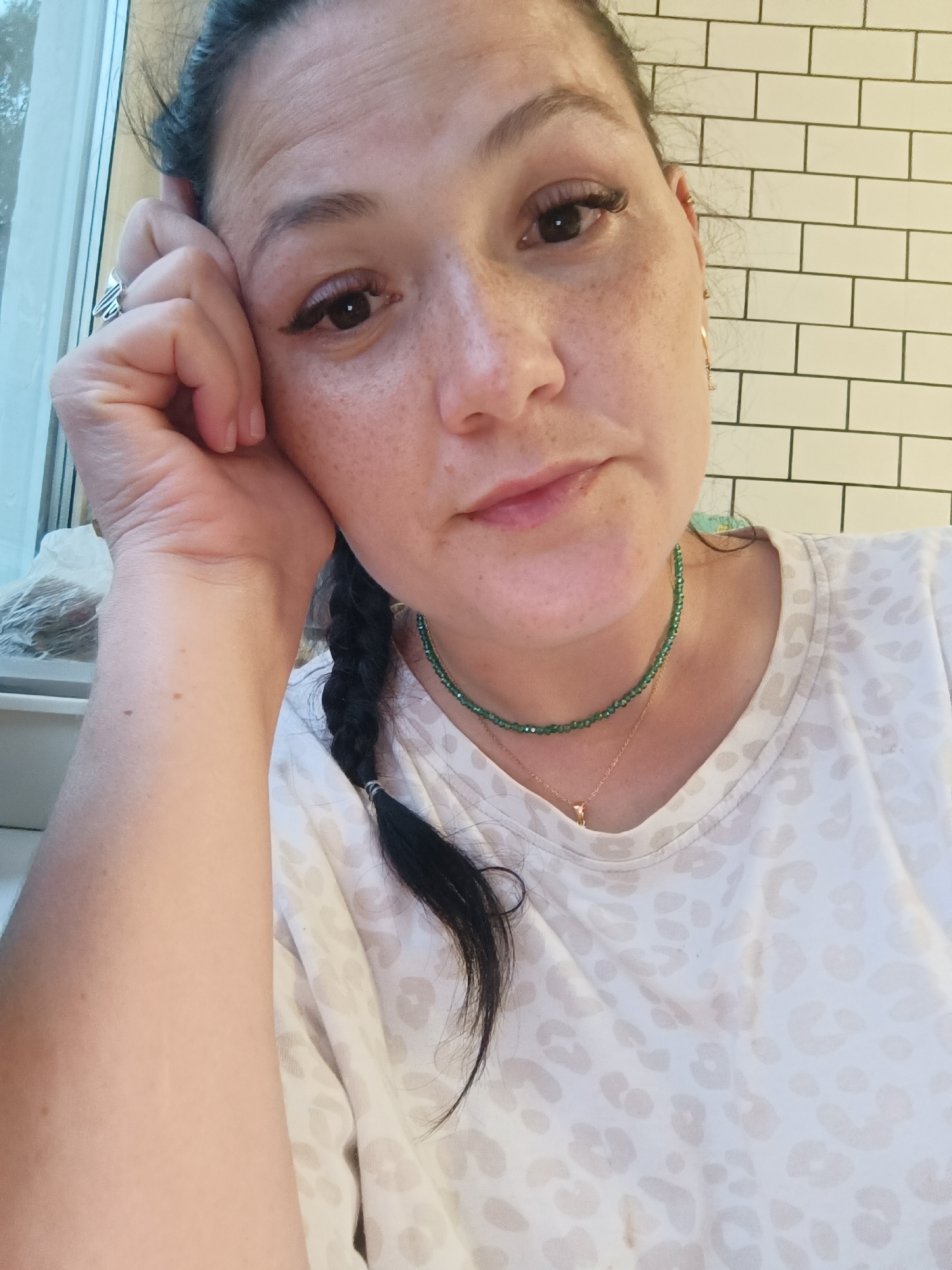 A medium shot photograph shows a woman resting her right hand on her cheek, her left arm is bent and her hand is resting against her face. She has dark hair pulled into a braid and is wearing a white t-shirt with a subtle leopard print pattern. She has a dark green beaded necklace and a delicate gold necklace with a small pendant. The background shows a white tiled wall and a window.