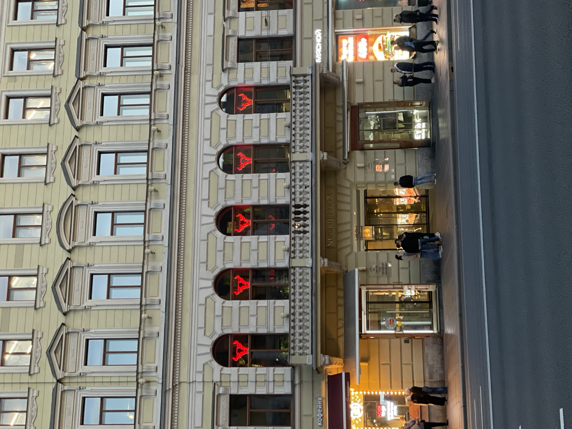 The image depicts an evening or nighttime street scene in Saint Petersburg, Russia, focusing on the ornate facade of a multi-story building. The building, rendered in a light yellow or cream color, features intricate architectural details. The upper floors display numerous rectangular windows, some adorned with decorative frames and triangular pediments, suggesting a classical architectural style.
On the lower floors, particularly prominent are several arched windows, each illuminated with a bright red neon sign resembling a bull or ram's head. These signs likely indicate a commercial establishment, possibly a restaurant or bar. Below these windows, multiple storefronts and entrances are brightly lit, contrasting with the dimming light of the evening.
Several pieces of text are visible on the building's facade and storefronts. In white Cyrillic letters, "МЯСНОЙ" (MYASNOY), meaning "MEAT" or "MEATY," is displayed above an entrance. Vertically oriented in Latin script, "NEVSKIJ PLAZA" is visible on a column, clearly identifying the building's location on or near Nevsky Prospekt. Further down, "КОФЕЙНА" (KOFEYNA), meaning "COFFEE SHOP," is visible on a maroon awning.
On the sidewalk in front of the building, several pedestrians are visible. Some individuals are walking, while others appear to be standing near the illuminated entrances, suggesting activity around the commercial establishments. The street itself is paved and appears empty of vehicles directly in front of the building, though it is clearly a bustling urban environment. The bright internal lights of the stores and the striking red neon signs are key indicators of the time of day.