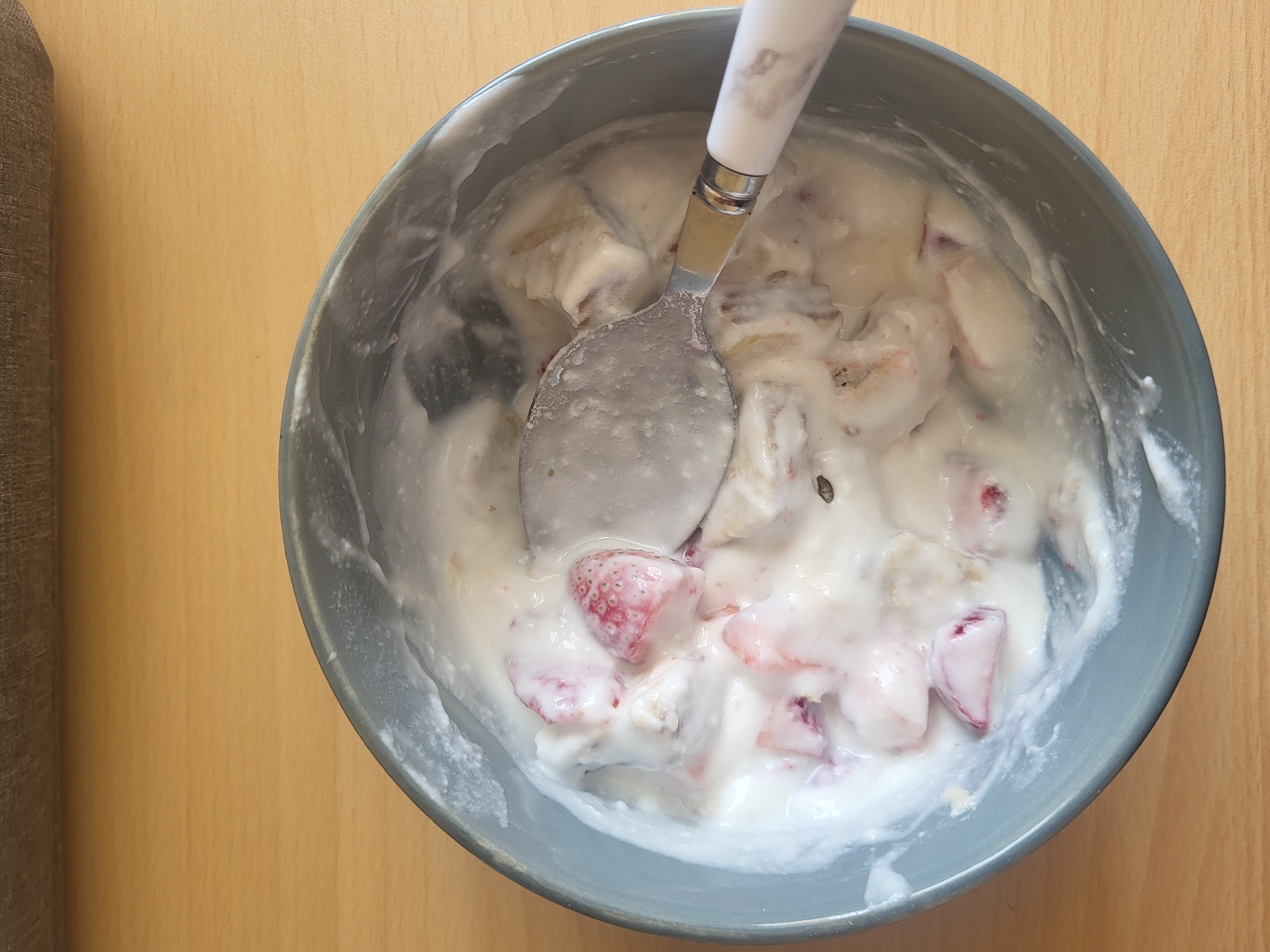 A top-down shot shows a gray bowl containing a yogurt mixture with sliced strawberries and bananas. A spoon with a white handle is in the bowl, partially submerged in the mixture. The bowl rests on a light brown wooden surface. A portion of a brown sofa or chair is visible on the left side of the frame. The location is unknown.
