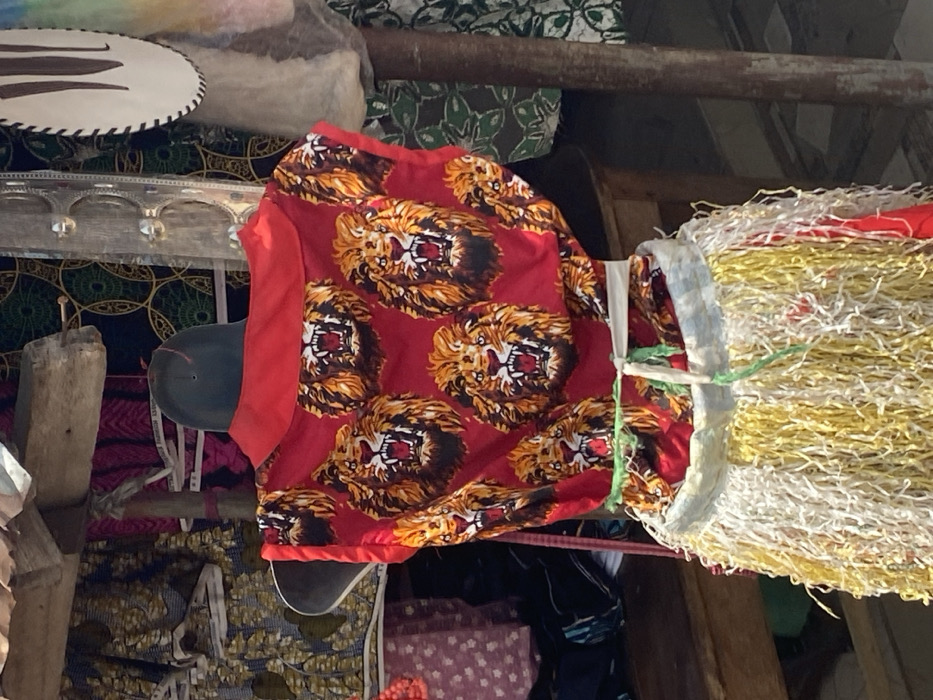 In an outdoor market stall in Afikpo, Nigeria, a headless torso mannequin is positioned centrally, facing away from the viewer. The mannequin displays a sleeveless red top featuring multiple repeating patterns of roaring lion heads and a red collar. Around the mannequin's waist, a skirt-like garment is tied with a green string; this garment is composed of dense, shredded strips of material, predominantly yellow and white, creating a fringed texture. Above the mannequin, a rough wooden beam of a stall structure is visible. In the background, various patterned fabrics are displayed, including dark fabric with light circular motifs and pink fabric. A weathered vertical wooden pole and a partially visible, irregularly shaped white object with a dark border are present on the right. A wooden bench or table is partially visible on the left. The scene is illuminated by daylight.