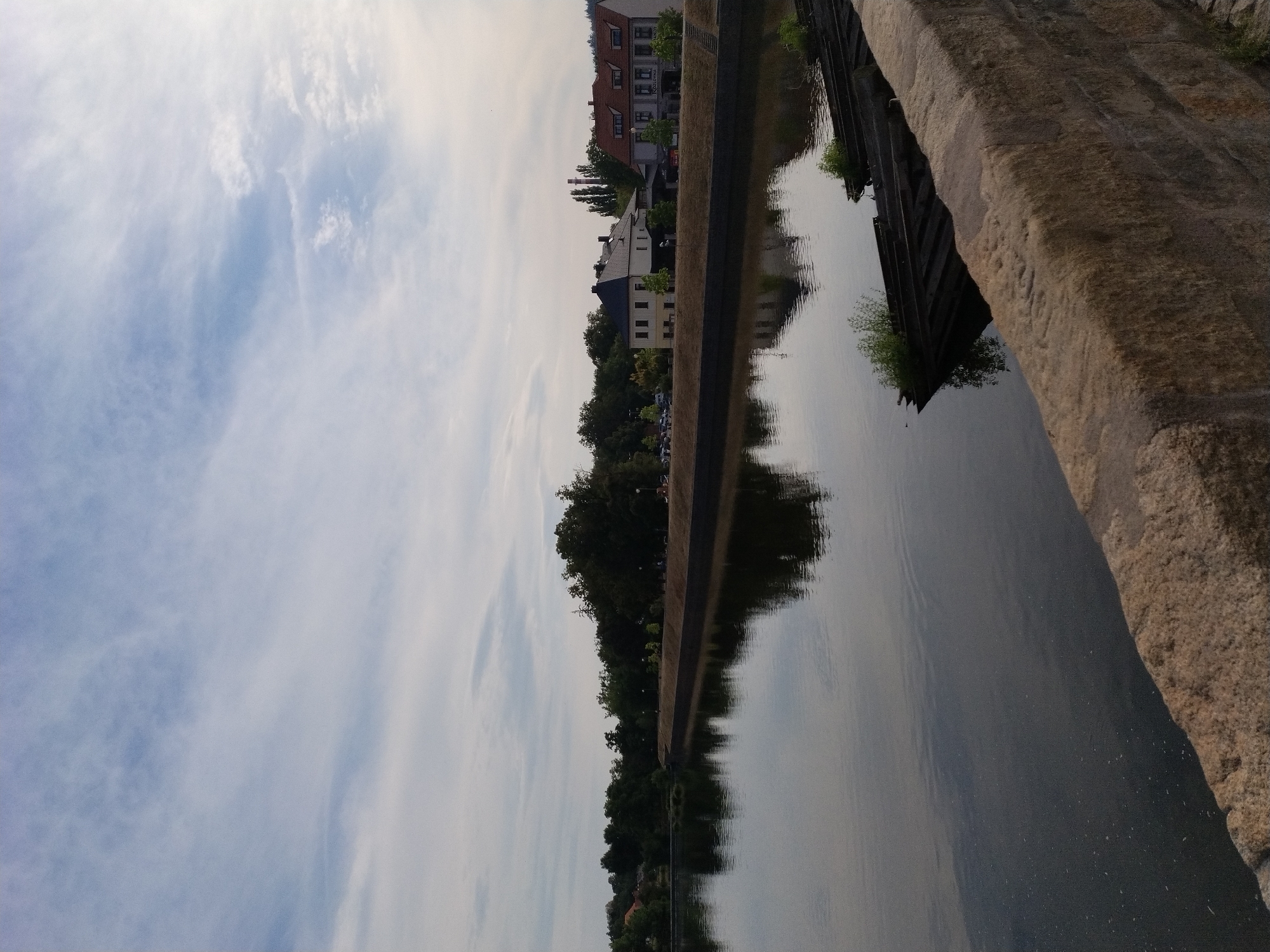 The image depicts a tranquil scene of the Otava River in Písek, Czechia. The river's surface is calm, reflecting the sky and the tree-lined bank on the opposite side. Several houses and buildings are visible along the riverfront, interspersed with lush green trees. In the foreground, a stone embankment or bridge railing made of rough-hewn blocks lines the lower portion of the frame. Partially submerged wooden structures, possibly old boats or pontoons, are visible in the water near the right edge of the image, with some vegetation growing on them. The sky is partly cloudy, with streaks of white clouds against a pale blue backdrop. The overall atmosphere is serene and picturesque, characteristic of a peaceful riverside setting in a town.