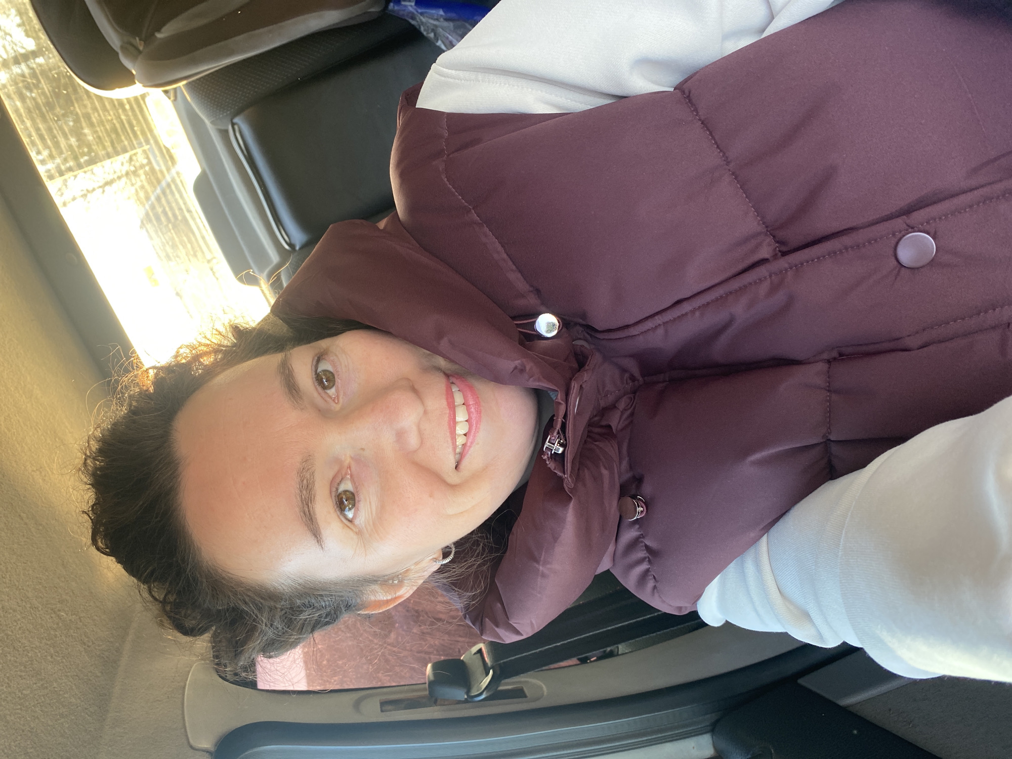A woman with curly brown hair and brown eyes is wearing a maroon puffy vest over a white long-sleeved shirt. She is smiling and facing the camera. She is inside a vehicle, possibly a car, with the interior visible in the background. Light is coming in from the left side of the frame, possibly through a window. The location is Прикамский, Russia.