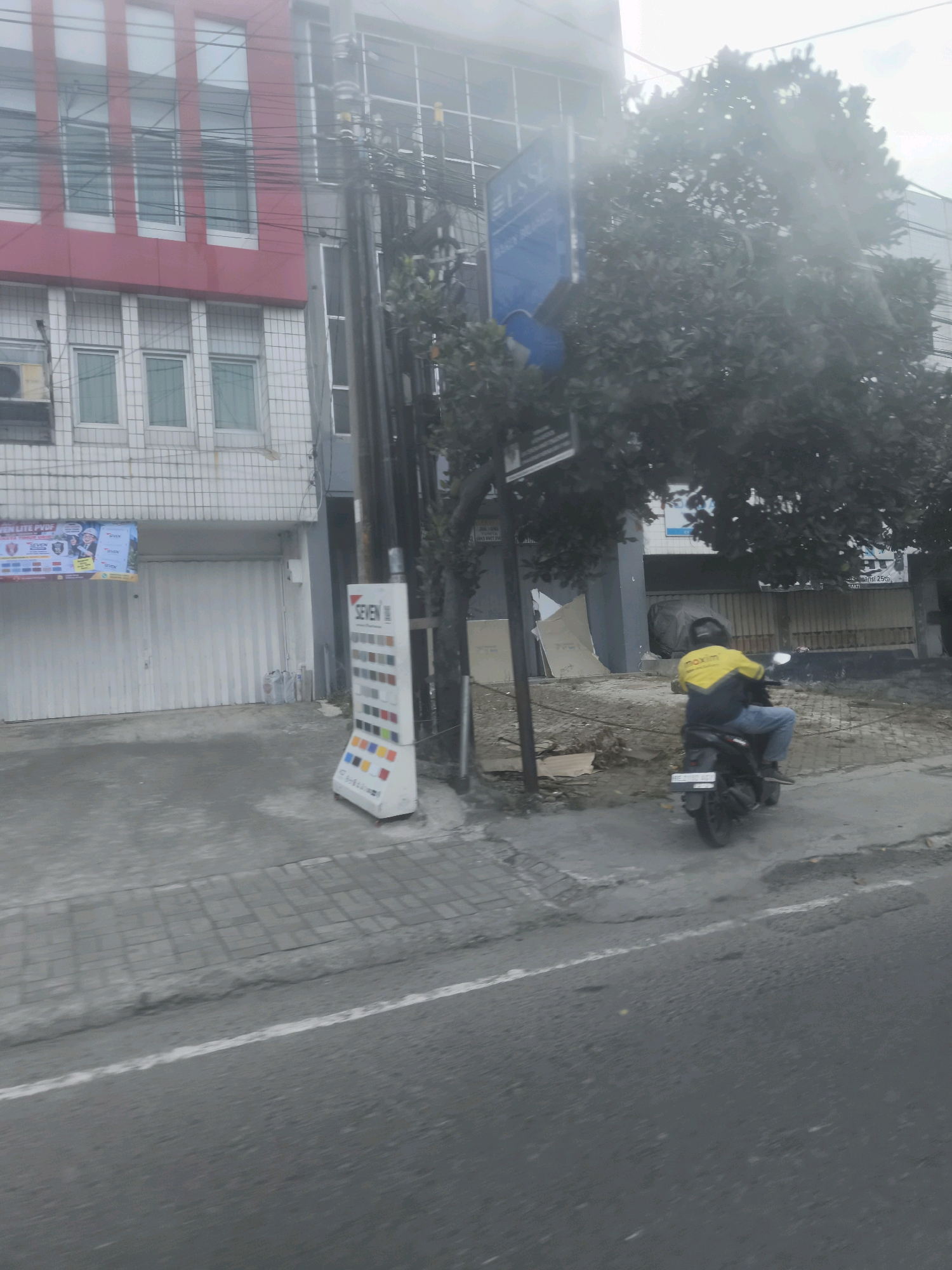 A motorcycle rider, wearing a yellow and black jacket and blue jeans, is observed from behind on a black motorcycle. The rider is moving away from the camera on a paved street in Bandar Lampung, Indonesia. In the background, a two-story building with a red and white facade is visible, featuring multiple windows. Adjacent to the building, a display stand with color swatches labeled "SEVEN" is positioned on the sidewalk. A large tree with dark green foliage partially obscures the scene. The sky is overcast, creating a muted lighting condition.