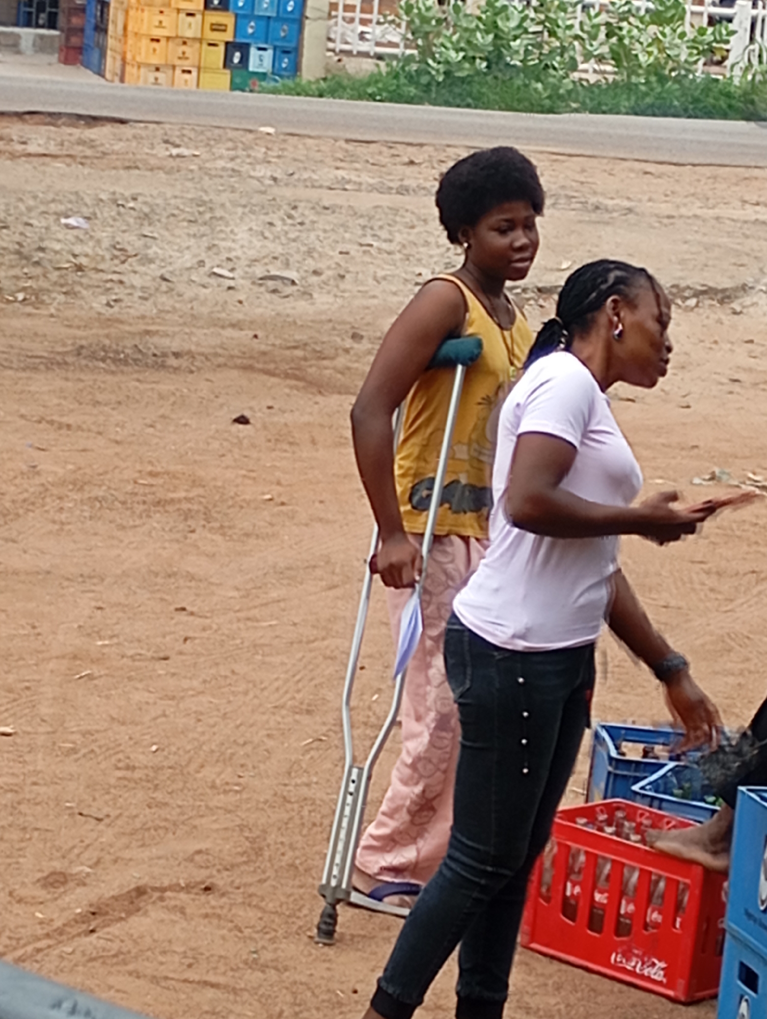 In Shendam, Nigeria, two women are seen standing on a dirt road. The woman on the left is using a crutch and wearing a yellow tank top and pink pants. She is looking to the right. The woman on the right, facing left, is wearing a light purple t-shirt and dark jeans. She is holding a small orange object in her right hand and looking towards the left. In the background, there are stacks of colorful crates, some containing bottles, and foliage.
