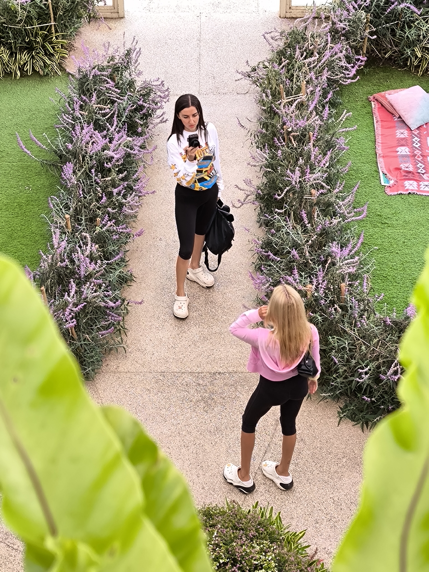 The image, taken from an elevated perspective, shows two women standing on a stone-paved path bordered by flowering plants with purple blossoms. The location is likely Candikuning, Indonesia based on the flora and possible Balinese-style architecture present.
One woman, with dark hair, wears a long-sleeved white top, black cycling shorts, and white clogs. She is holding and looking at a camera. She also has a black shoulder bag.
The other woman, with long blonde hair, is wearing a light pink hoodie, black cycling shorts, white clogs with black socks, and a small black purse over her shoulder. She is facing away from the camera and appears to be adjusting her collar or hair.
On one side of the path, a section of green turf is visible with a patterned rug and pillows on it. The image appears to be taken outdoors during daylight. A large green plant with large leaves is partially visible in the foreground of the image, obstructing the view. The flowering plants on either side of the path appear to be arranged to create a sort of archway. The background is slightly blurred, but appears to be a wall or structure.