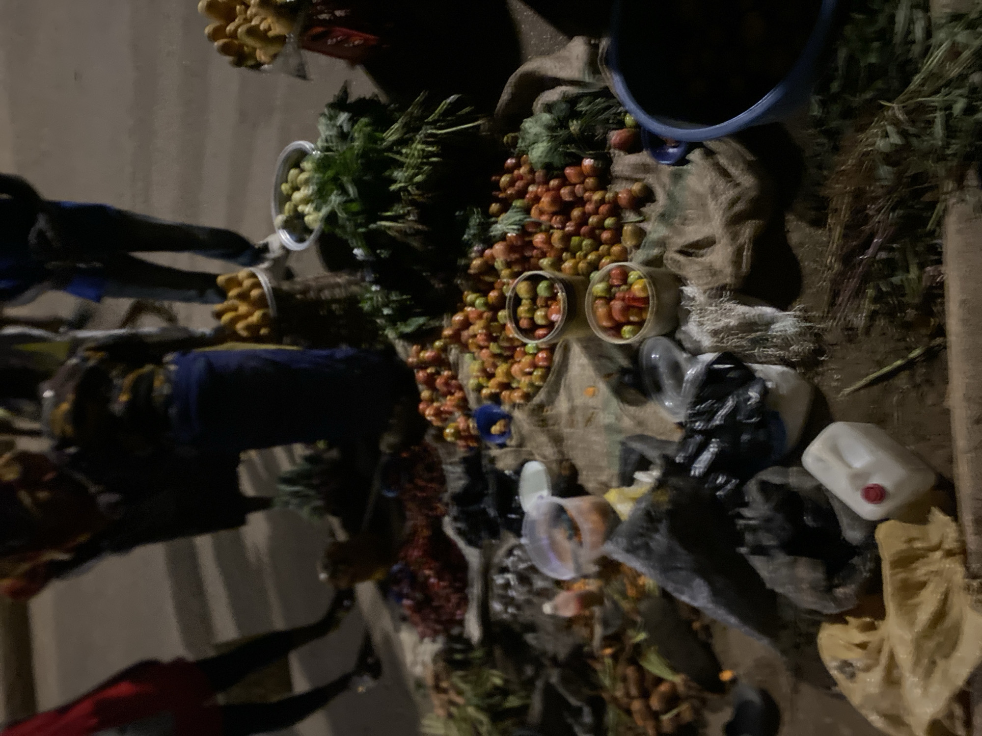 This media file captures a dimly lit outdoor market scene in Bukuru, Nigeria, likely during the night or early morning hours. The setting shows various fresh produce laid out on the ground or low surfaces, predominantly on burlap sacks.
The central focus is on an abundance of small, round fruits and vegetables, primarily tomatoes, displayed in large piles and contained within clear plastic buckets. These items exhibit a range of colors from reddish-orange to greenish-yellow. Adjacent to them are bundles of green leafy vegetables, also placed in clear plastic containers, suggesting they are ready for sale. Further produce includes yellowish-brown, irregularly shaped items, possibly yams or cassava, seen in white buckets and scattered piles. Towards the bottom right, elongated root vegetables, resembling carrots or sweet potatoes, are visible.
Several objects are scattered around the market stall: a large blue plastic bucket is partially visible at the top right, while a white plastic jerrycan with a red cap and multiple black plastic bags are positioned on the right side.
Several people are discernible in the image, though their figures are somewhat indistinct due to the low light and motion blur. On the left side, a person wearing a blue top is visible, possibly attending to the stall. Another individual in a red shirt is partially seen at the bottom left. The overall activity depicted is that of commerce in a local market, with vendors displaying goods and potential customers browsing or making purchases. There is no visible text.