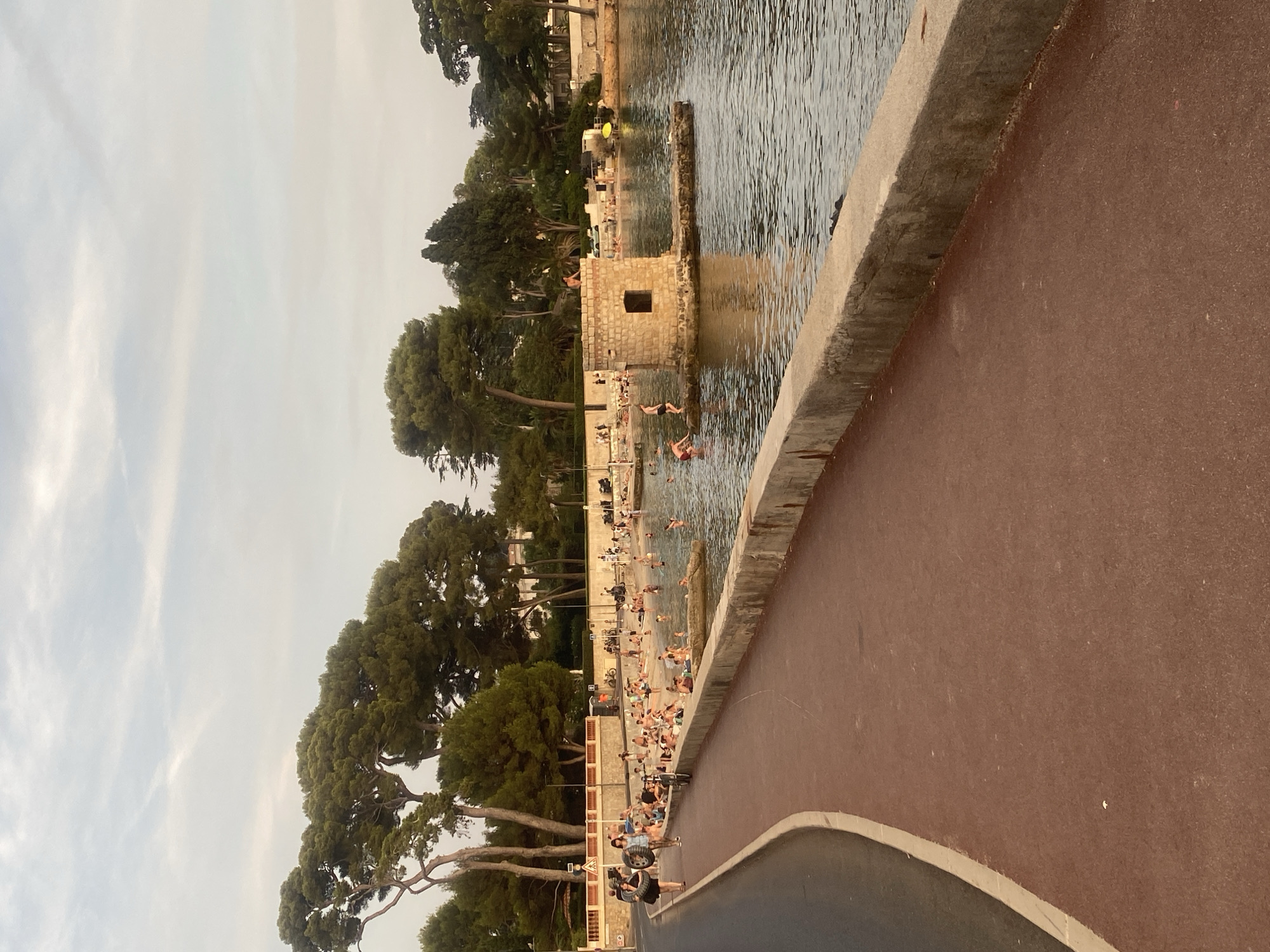 The image depicts a view of the waterfront in Antibes, France, likely taken from a paved walkway. The walkway, surfaced with red asphalt and bordered by a light-colored curb, curves along the left side of the frame, leading towards the water. A stone retaining wall separates the walkway from a sandy beach area crowded with people. Many individuals are sunbathing, sitting, and wading in the clear, calm water.
In the water, slightly offshore, is a small, square stone tower built on a rocky platform. One person is visible standing atop this tower. The water extends to the right, where more of the coastline with trees and buildings can be seen. Tall pine trees line the upper edge of the beach and extend into the background, with a residential building partially visible through the foliage. The sky is overcast with soft, diffused light. On the walkway in the foreground, two women are walking away from the camera, one carrying what appears to be a spare tire. Several bicycles are also present near the beach entrance. The scene conveys a relaxed atmosphere of a popular summer recreational area.