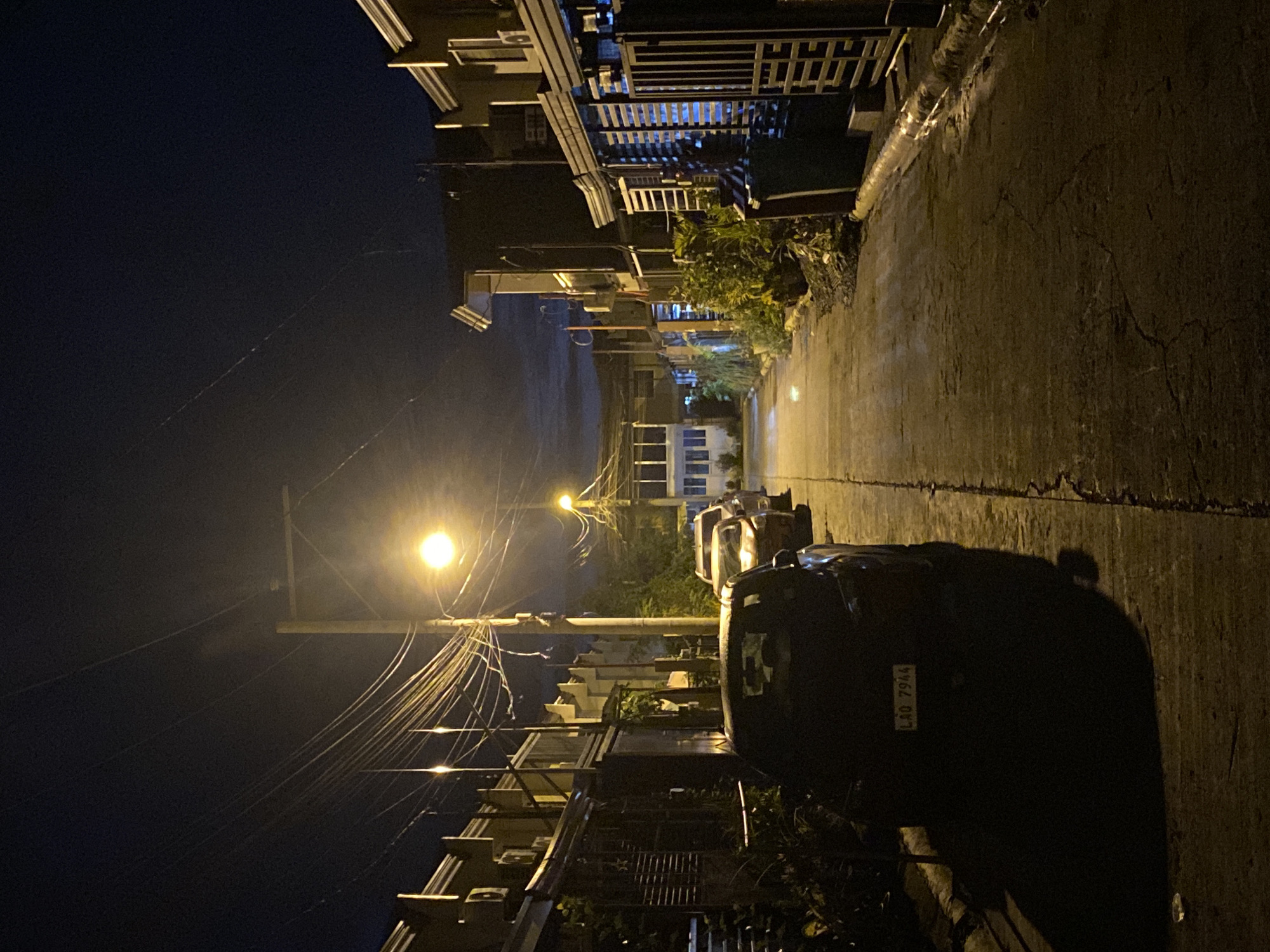 A nighttime street scene in a residential area of Davao City, Philippines, is depicted. The street is lined with houses and parked cars, illuminated by a street light and the lights from the houses. The sky is dark, with a few wisps of clouds visible. The pavement is wet, suggesting recent rain. A car with license plate "LAQ 7944" is parked in the foreground. The overall impression is one of quiet and stillness.