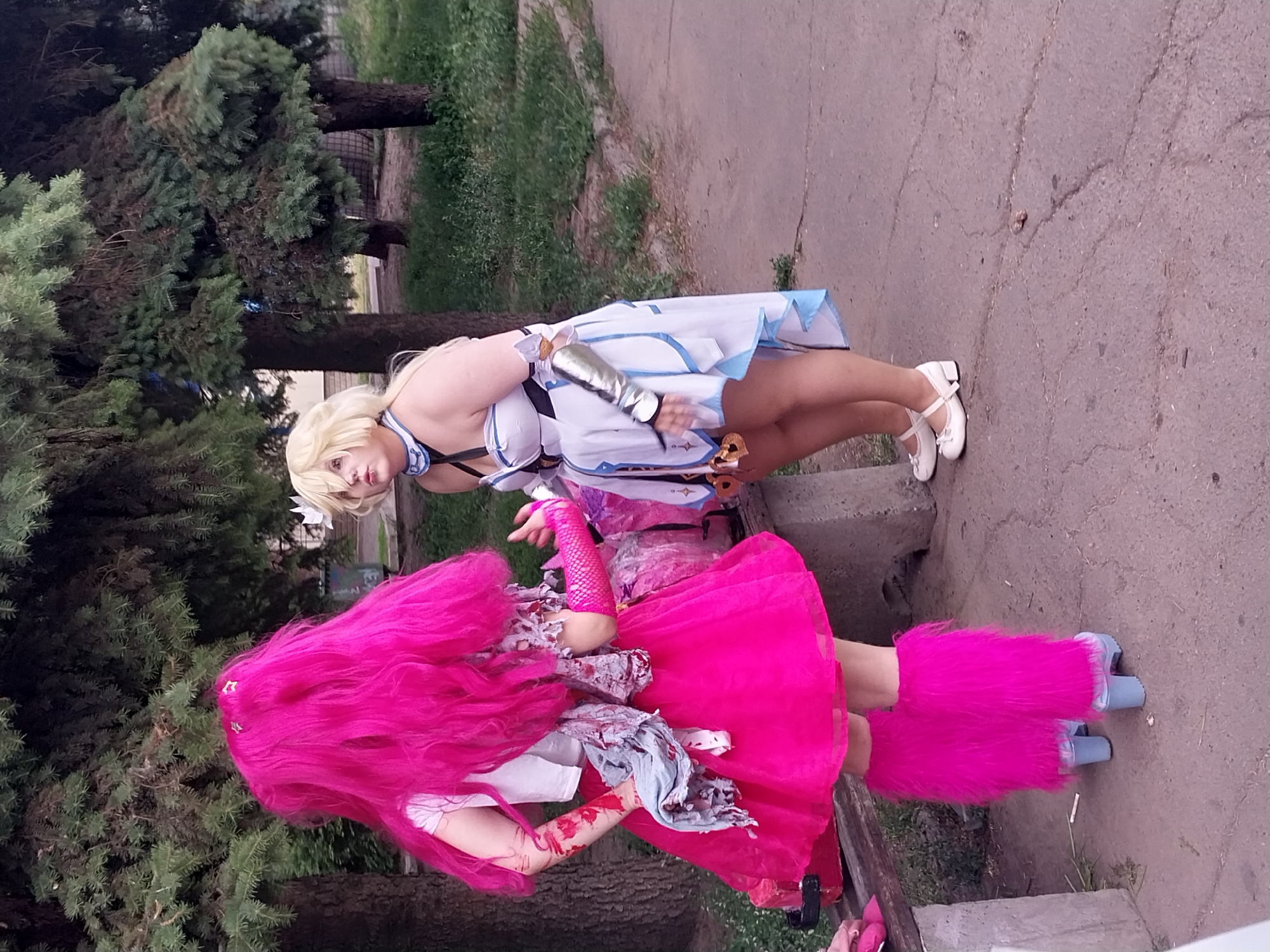 This media file features two individuals in cosplay, posing outdoors. The subject in the foreground has long, vibrant pink hair and is wearing a bright pink dress and furry pink leg warmers over blue platform shoes. Their arms appear to be adorned with simulated blood and tattered fabric, suggesting a zombie or horror-themed character. The second subject, in the background, has blonde hair and is dressed in a white and blue outfit with silver arm armor. They are wearing white Mary Jane shoes. Both subjects are standing on a cracked concrete path, with lush green trees and grass surrounding them. The lighting suggests it is daytime, likely overcast as there are no strong shadows. The overall scene conveys a playful, costume-event atmosphere, possibly a cosplay gathering or photoshoot. There is no visible text in the image.