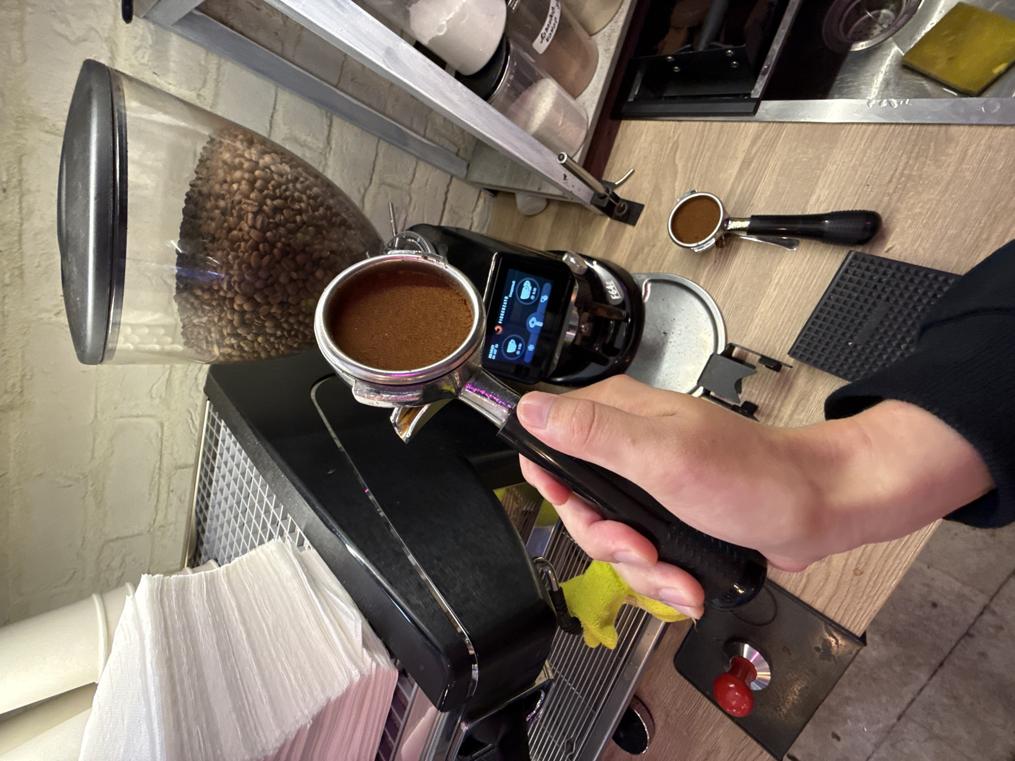 The image depicts a close-up of a person's hands preparing to make an espresso, likely in a cafe or a home barista setup. The dominant subject is a portafilter filled with finely ground coffee, held by a hand with short fingernails. The portafilter is positioned over the drip tray of a coffee machine. In the background, a coffee grinder with a large hopper filled with coffee beans is visible, along with a stack of paper cups and a digital display on the coffee machine showing brewing parameters. Other objects include a tamping mat, a tamper, and various containers likely holding coffee-related supplies. The scene is set on a wooden countertop, with a brick wall in the background. There is no discernible indication of the specific location being Moscow, Russia, as the image lacks any geographical markers or recognizable landmarks.