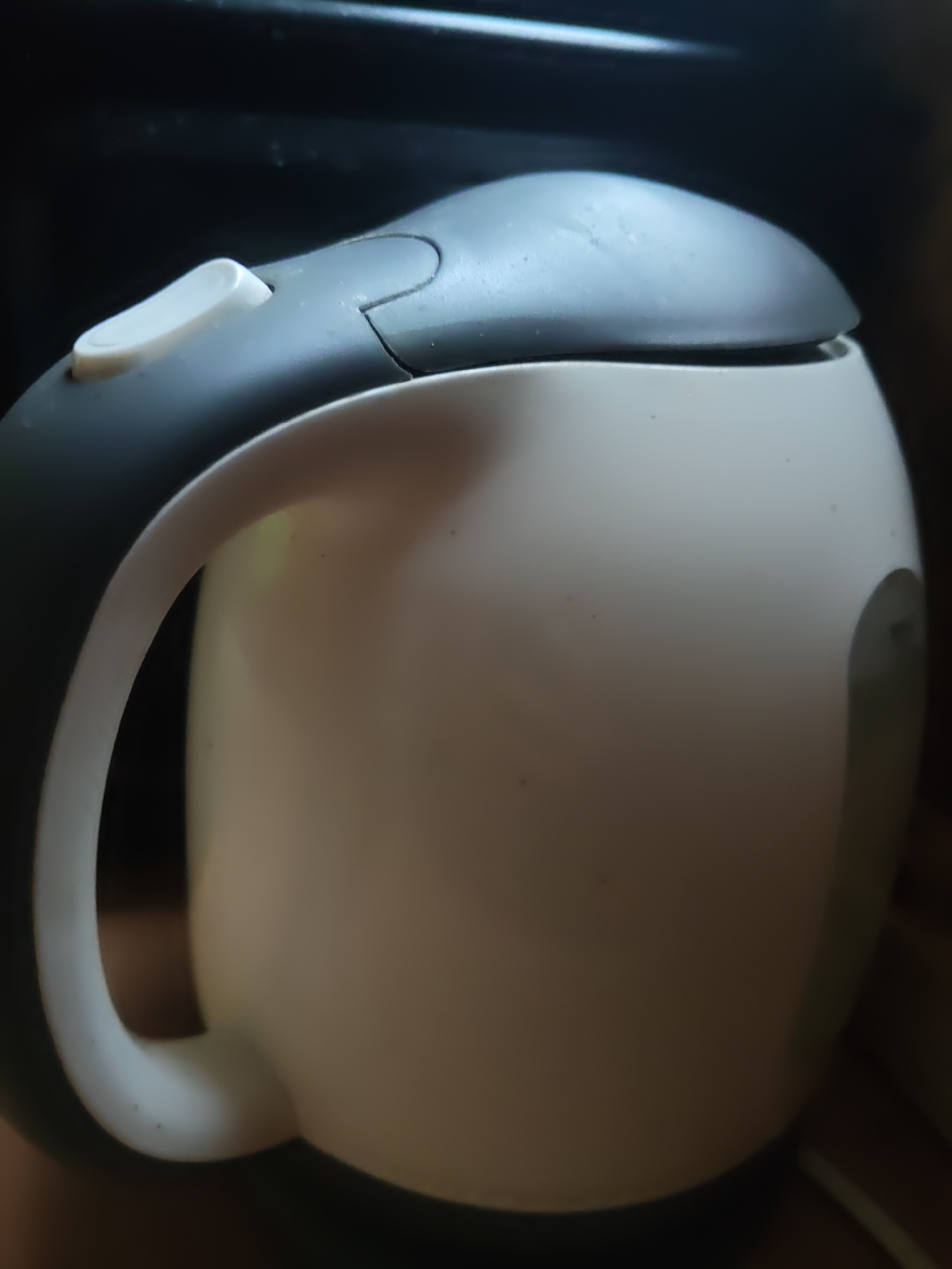 An electric kettle, predominantly white with a dark gray lid and handle, is positioned in a slightly angled, close-up shot. The kettle features a prominent white button on the lid, indicative of its on/off switch. The surface of the kettle appears matte and shows minor signs of wear or dust. The background is dark and out of focus, offering no discernible details of its surroundings. There are no people or animals present in the image. The lighting is dim, creating shadows and highlighting the textured surfaces of the kettle. The overall scene suggests an indoor setting, though the specific location is not identifiable.