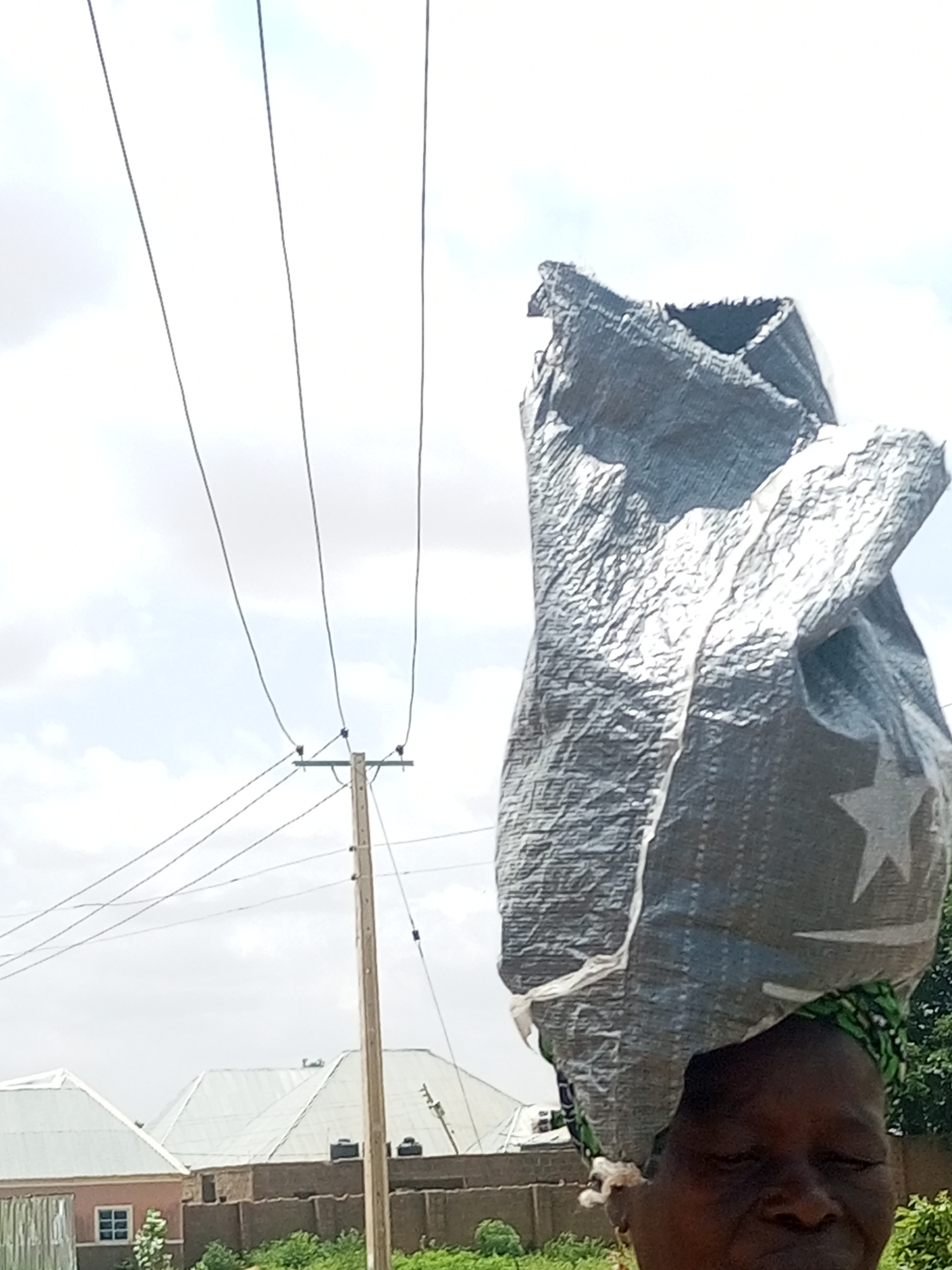 An African woman with dark skin, wearing a green and white patterned headwrap, carries a large, silver, tarp-like bag on her head. The bag is creased and appears to contain a bulky item, with a white star visible on its surface. Her eyes are closed, and her face is partially visible. The background shows a utility pole with multiple wires, a partially obscured building with a light pink facade and a white roof, other buildings with metal roofs, and lush green vegetation. The scene is set outdoors under a cloudy sky, possibly in Tempure, Nigeria.
