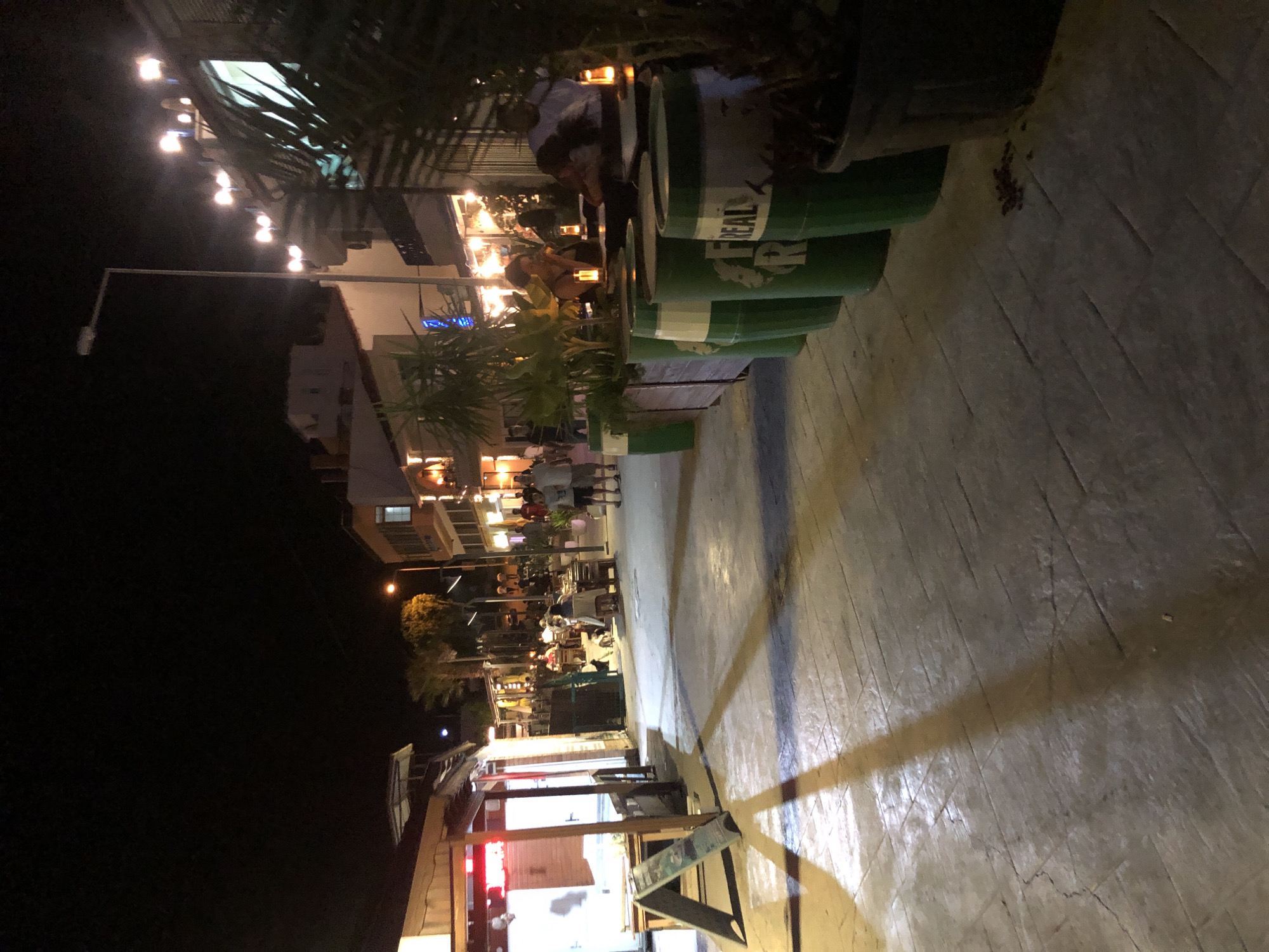 The image captures a nighttime scene on a street in Karaburun, Turkey, likely a commercial area with restaurants or cafes. The foreground features a paved walkway with a shadow cast from a structure or object on the left. Several large green barrels, possibly used as planters or decorative elements, are arranged in a line along the right side of the walkway. Some of these barrels have white lettering, including the word "REAL" visible on one. In the middle ground, tables and chairs are set up, suggesting an outdoor dining area. People are seated at these tables, engaged in conversations or dining. Behind the tables, buildings with illuminated windows and external lights line the street, indicating shops or other establishments. A streetlamp is visible in the distance, illuminating the scene. The overall atmosphere is one of a lively evening in a coastal town.