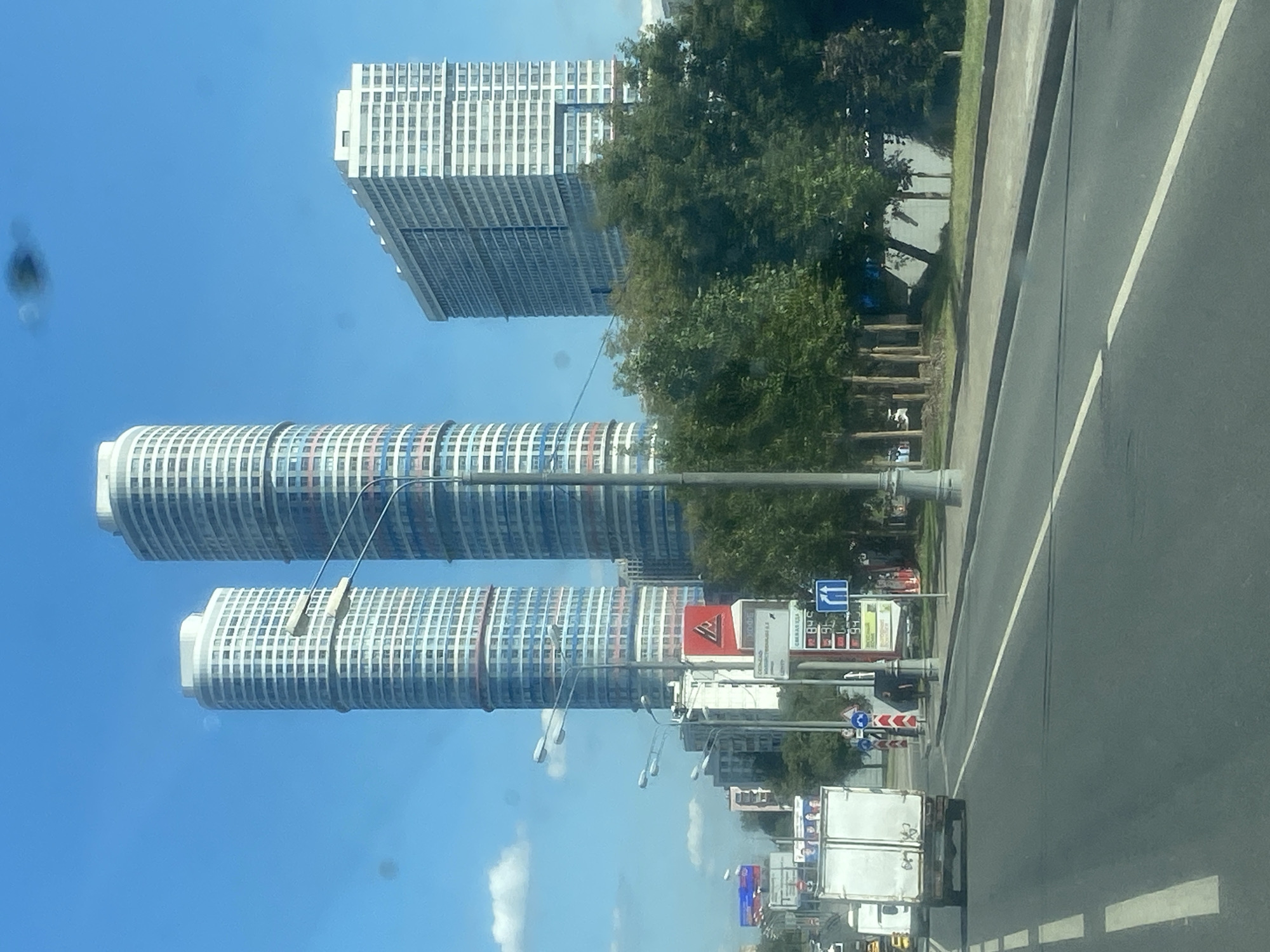 A daytime exterior shot captures a city street scene in Unknown, Unknown. In the background, three modern skyscrapers dominate the skyline. The two cylindrical towers on the left are white with horizontal bands of red and blue, featuring numerous windows. The building on the right is rectangular and also white with many windows. In the midground, a line of lush green trees separates the street from the buildings. A white delivery truck is visible on the road, moving away from the camera. Various street signs and lampposts are present along the side of the road. The sky is a clear blue with a few wispy white clouds.