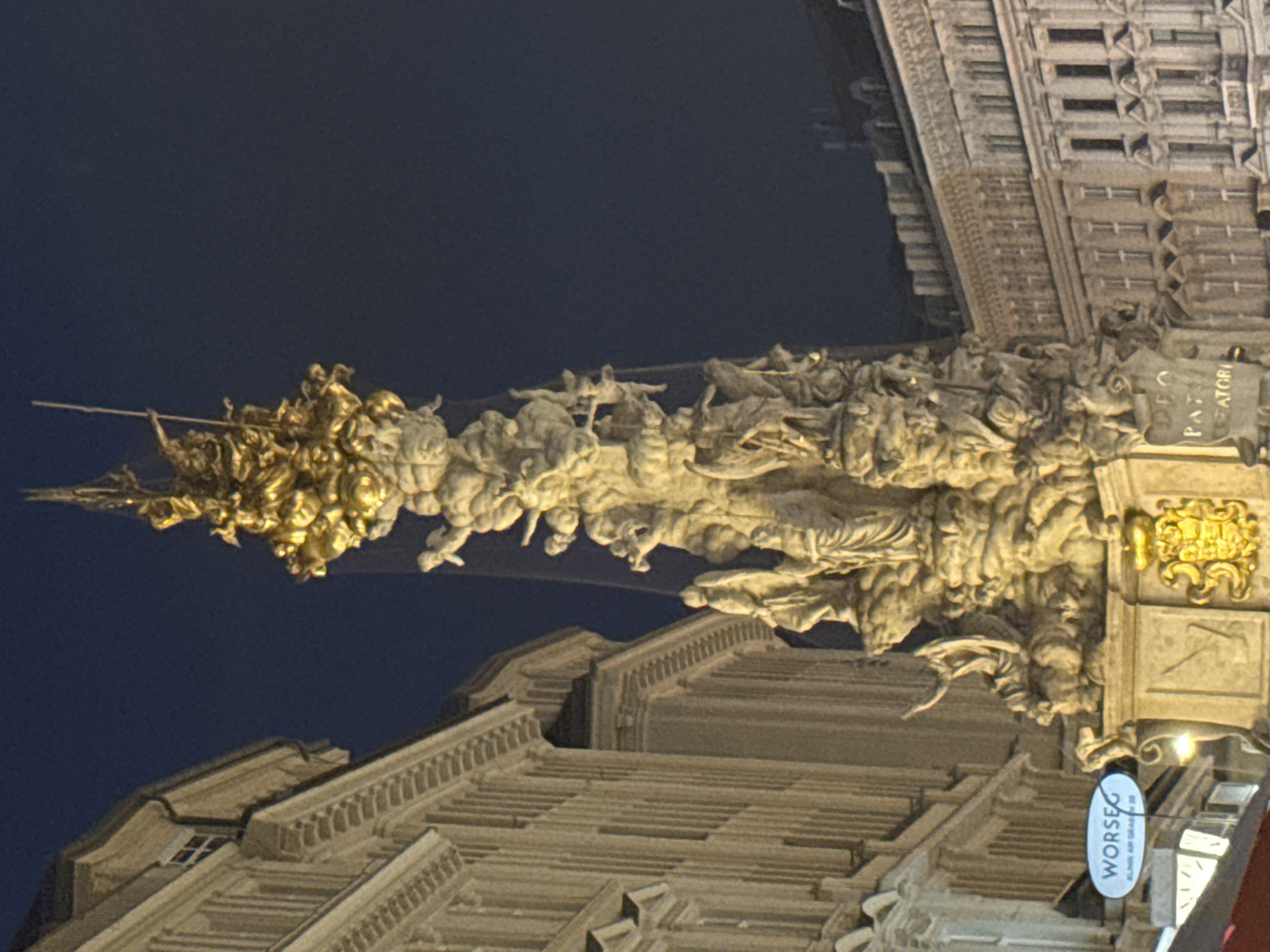 The image shows the Pestsäule (Plague Column) in Vienna, Austria, at night. The column is a baroque monument made of marble and gold, depicting a scene of the Holy Trinity and various figures from Christian iconography, as well as allegorical representations of the fight against the plague. It is located on the Graben street in Vienna's first district. The monument is illuminated by artificial lights, which create a dramatic effect against the dark night sky. The surrounding buildings are also visible, with their facades lit up. A sign for "WÖRSEG" is visible on the left side of the image, indicating a nearby establishment. The image is taken from a low angle, emphasizing the height and grandeur of the Pestsäule.