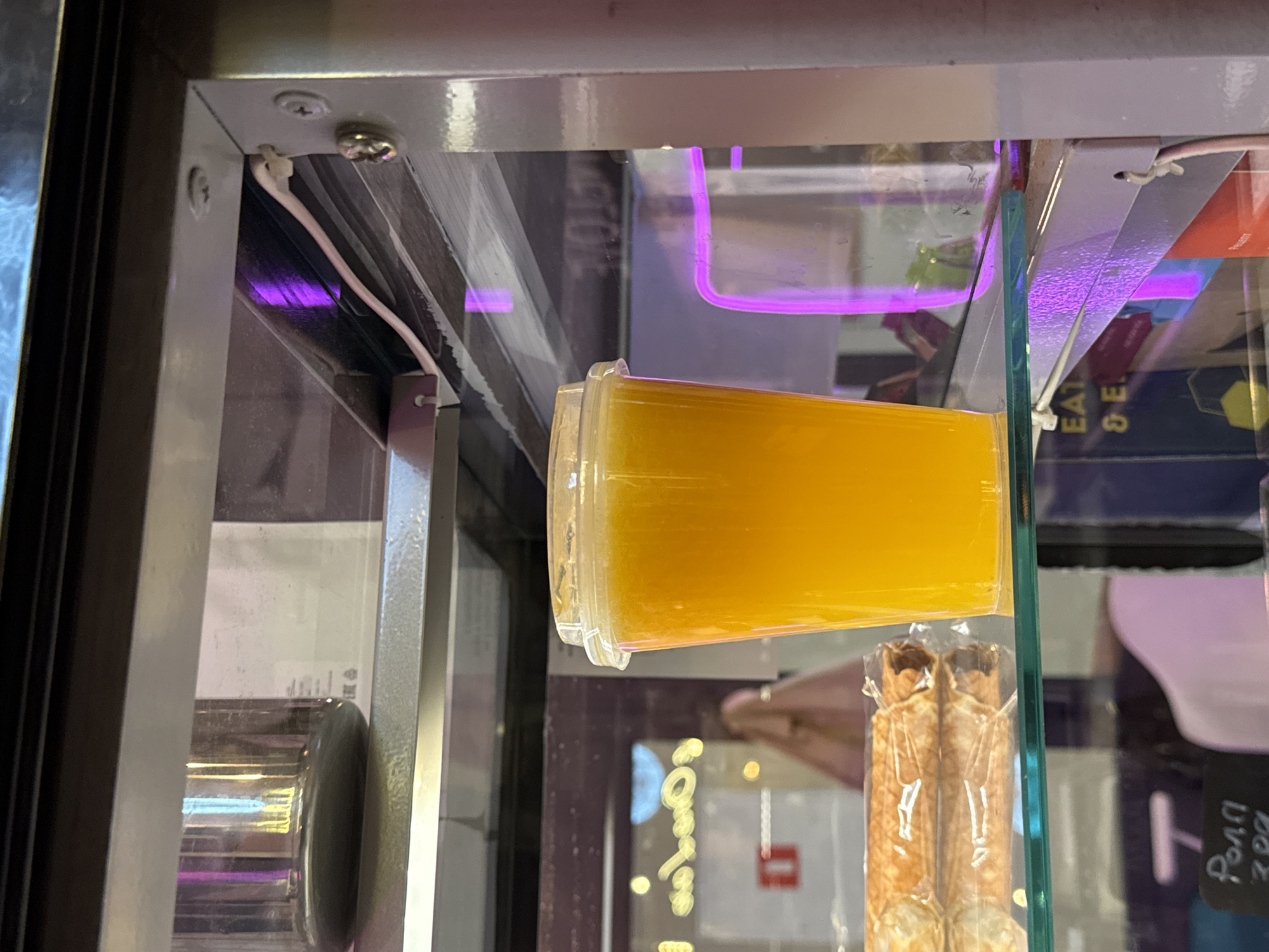 A close-up shot shows a clear plastic cup filled with bright orange liquid, likely juice, sitting on a glass shelf. The cup has a transparent lid. To the left of the cup, a rolled food item, possibly a sweet pastry or dessert, is visible, wrapped in clear plastic. The background suggests an indoor environment, possibly a cafe or shop, with shelves and display cases. Reflections on the glass shelf indicate lights and possibly other items. The overall lighting appears to be a mix of natural and artificial, with purple hues suggesting neon or colored lighting from the display. The scene is static with no people actively interacting with the displayed items. The location is identified as Moscow, Russia.