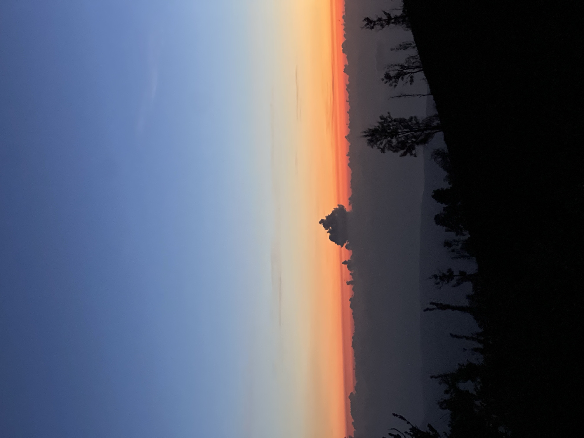 The image depicts a sunset or sunrise over what appears to be mountainous terrain. The sky transitions from blue on the left to orange and red towards the horizon line, where there are visible clouds. Silhouetted trees and vegetation are present on the right side of the image, suggesting a landscape with plant life. The location is Unknown, Indonesia, but no specific landmarks or clues within the image confirm the city. No people are visible in the image.