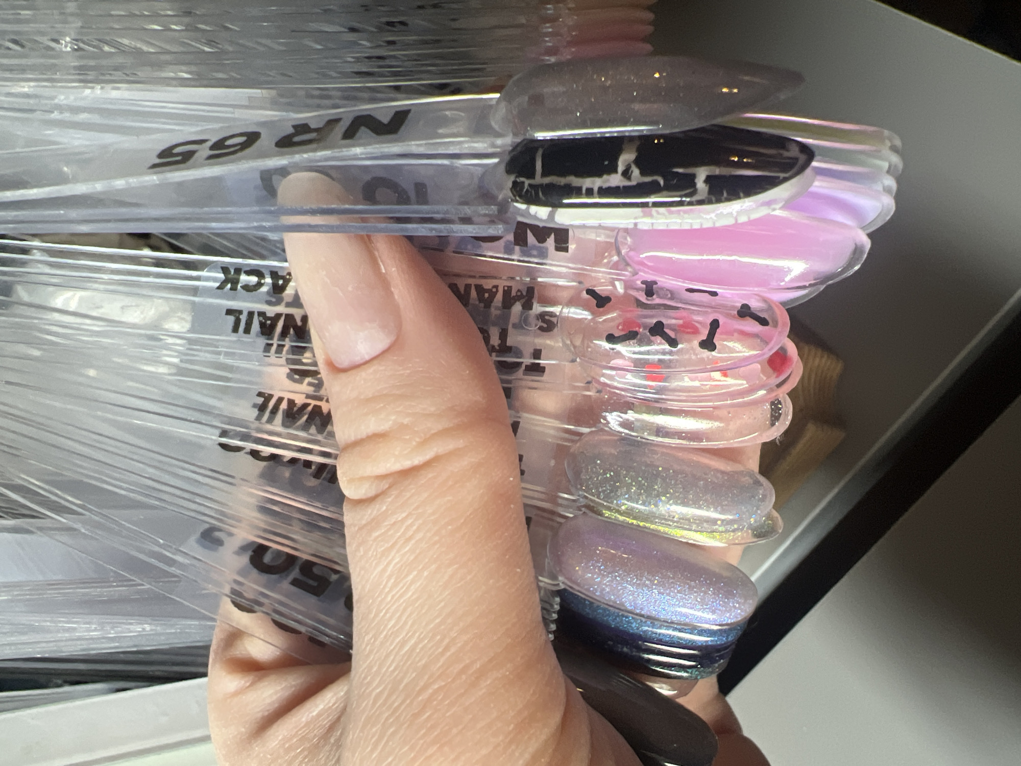 A close-up shot captures a human hand holding a fan of clear plastic nail polish swatch sticks, showcasing a variety of nail art and color samples. The hand, with visible skin texture, supports the collection of sticks, which are layered and slightly fanned out.
The tips of the swatch sticks feature diverse nail polish designs: a light gray with glitter, a black polish with a distinctive white marble or crackle effect, various iridescent and shimmery pinks and purples, a clear stick embedded with small black and red shapes (possibly tiny bones or hearts), and a vibrant glittery blue-to-purple gradient.
Visible black text on some of the clear sticks, often upside down or partially obscured, includes "NR 65", "NACK" (likely part of "BLACK"), "NAIL", "MAN", "TO...", and "50 S". The background is dark and blurred, suggesting an indoor setting, possibly a nail salon or personal collection, under artificial lighting that highlights the glossy and glittery finishes of the polishes. The activity depicted is the presentation or examination of these nail polish samples. This scene takes place in Tbilisi, Georgia.