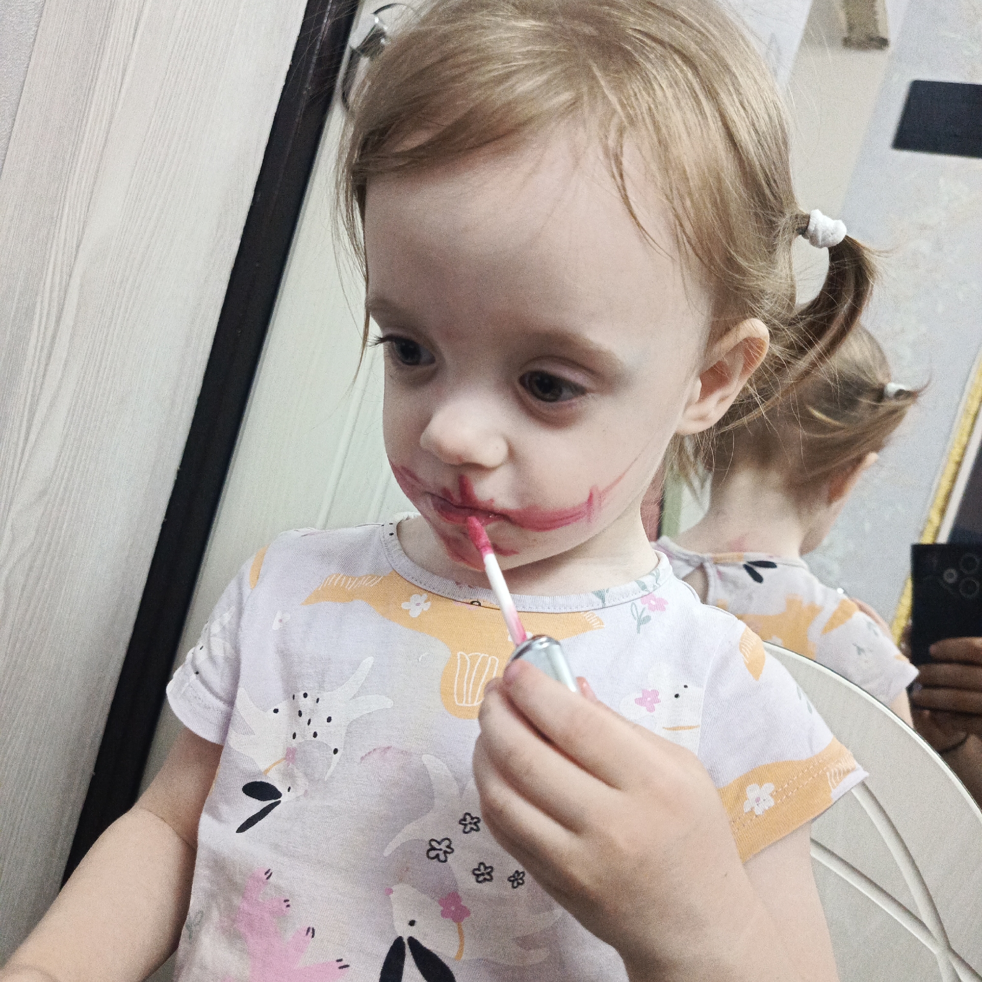 A young, fair-skinned child with light brown hair styled in two small ponytails is applying red lip gloss. The application is imprecise, with streaks of color around the mouth. The child is wearing a light-colored patterned shirt. A mirror reflects the child and someone holding a smartphone, partially visible. The location is identified as Марковский, Russia. The setting seems to be indoors, possibly a domestic environment.