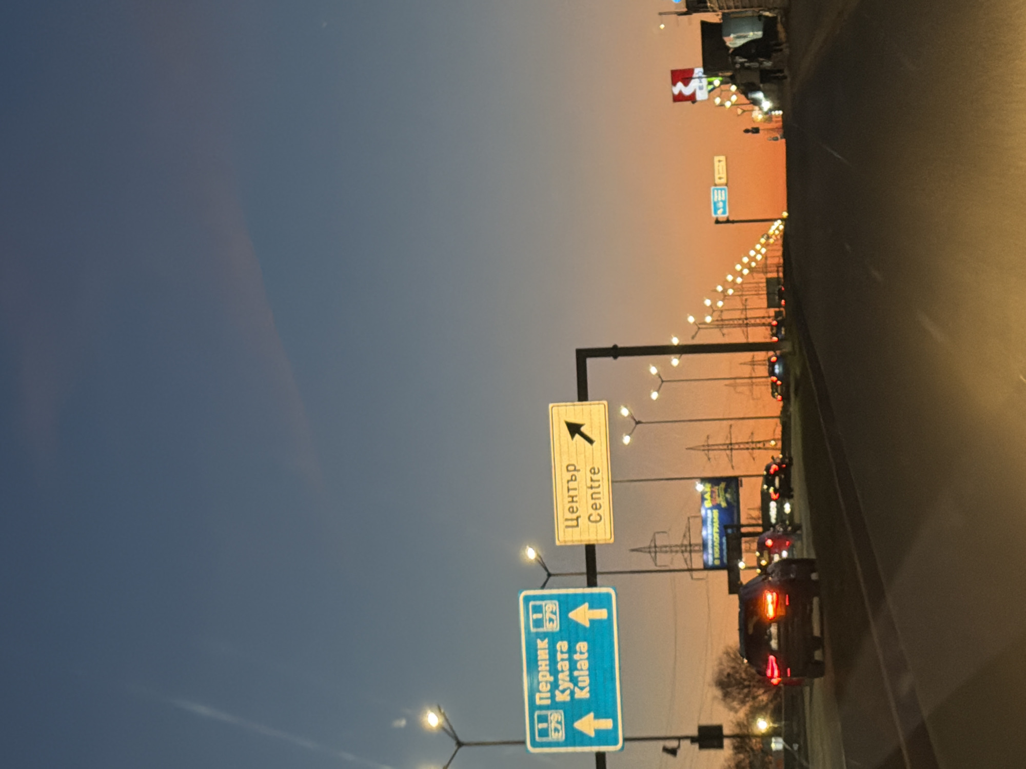 The image depicts a roadway in Sofia, Bulgaria at dusk. A blue and white directional road sign indicates the direction to Pernik and Kulata. A yellow sign directs toward the "Centre". Cars drive along the roadway. Streetlights illuminate the road. The sky is a gradient of blue to orange, likely indicating sunset or sunrise.