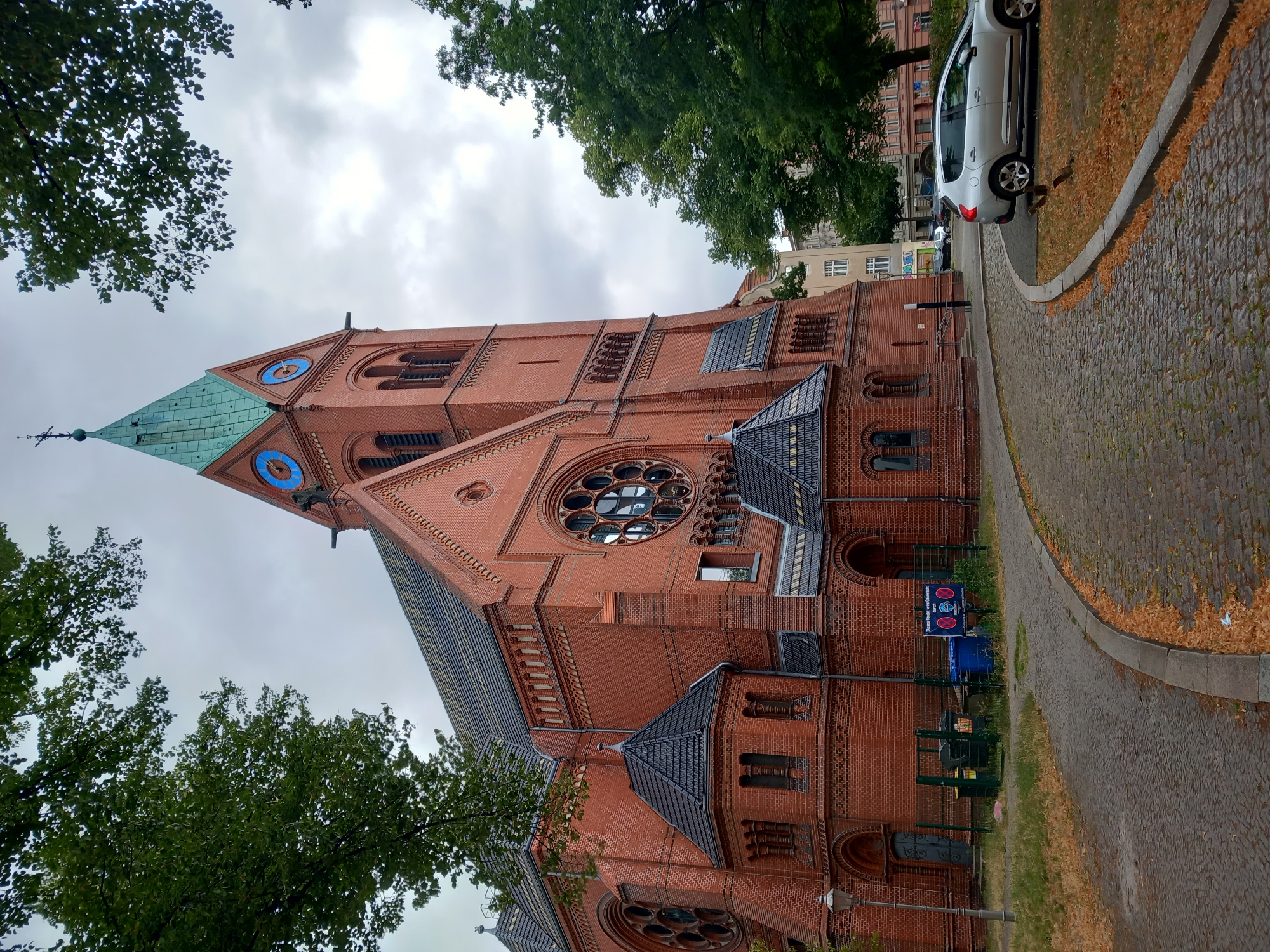 A brick church with a green spire and blue clock faces stands against a cloudy sky. The church is surrounded by trees, and a cobblestone path curves around it. A silver car is parked on the side of the path. The image appears to be taken in Berlin, Germany, as suggested by the architecture and the presence of a German sign in the background. The scene is peaceful and quiet, with no people or animals visible. The overall impression is one of a serene and historic urban setting.