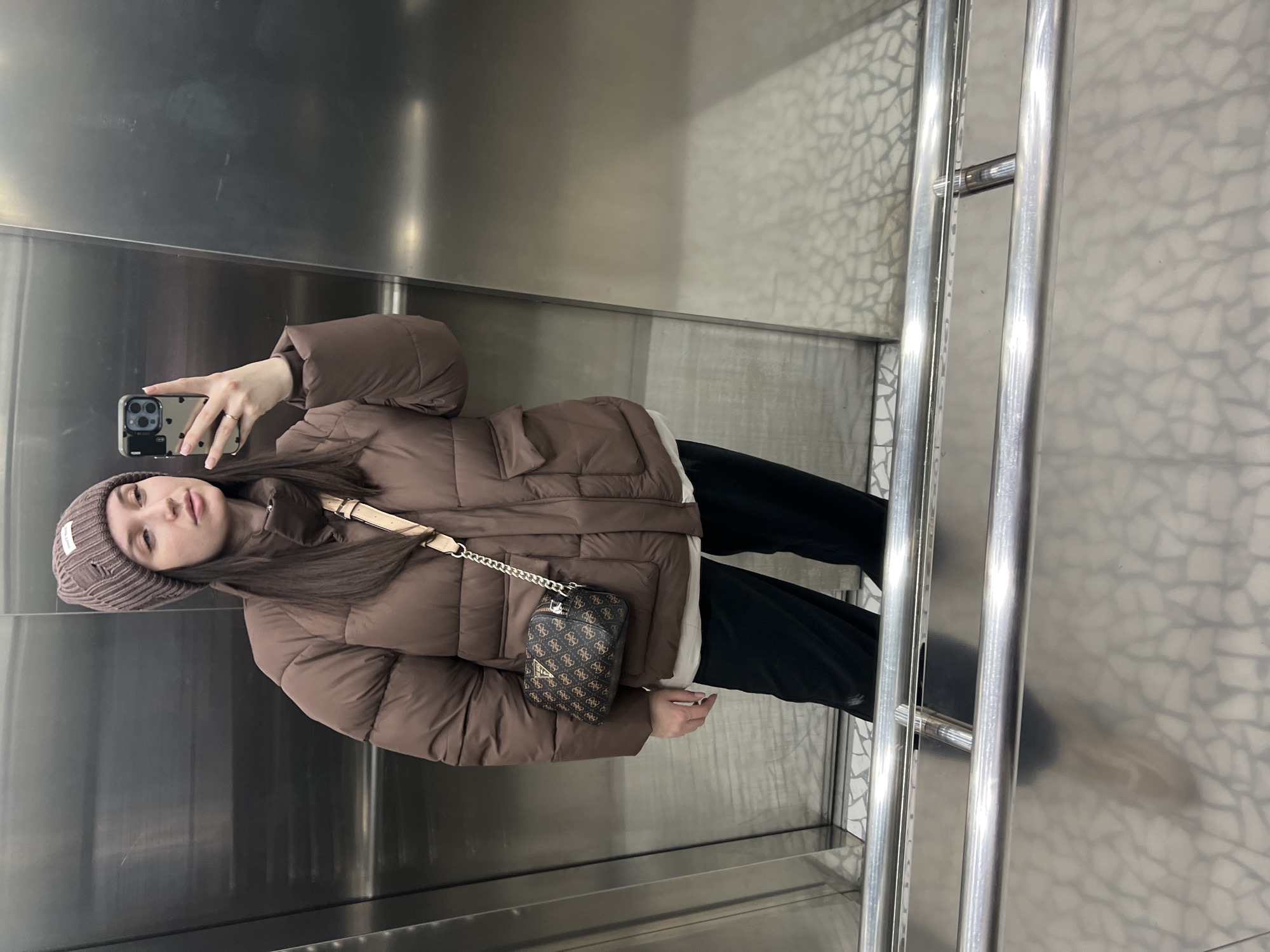 A woman is taking a selfie in an elevator. She is wearing a brown puffer jacket, a brown beanie, and dark pants. She has a small purse hanging from her shoulder. The elevator has metal walls and a patterned floor. The location is likely Tyumen, Russia, based on the image's origin.